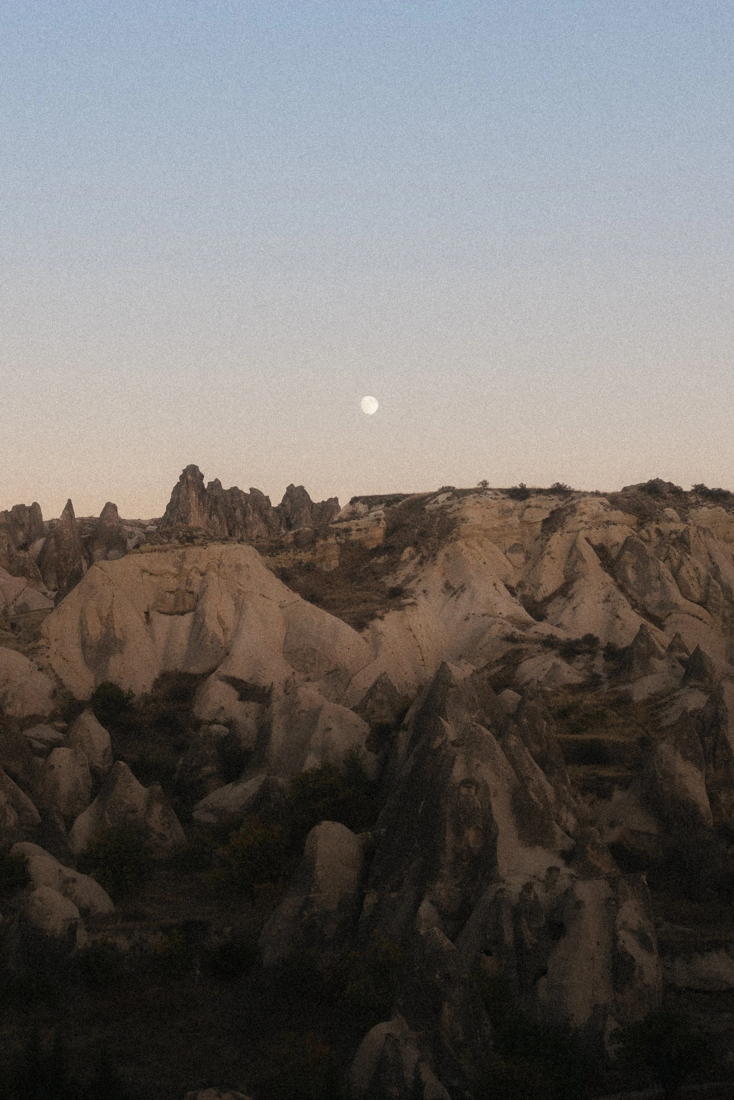 A rocky landscape during twilight or dawn, with a visible moon in the sky above the horizon.