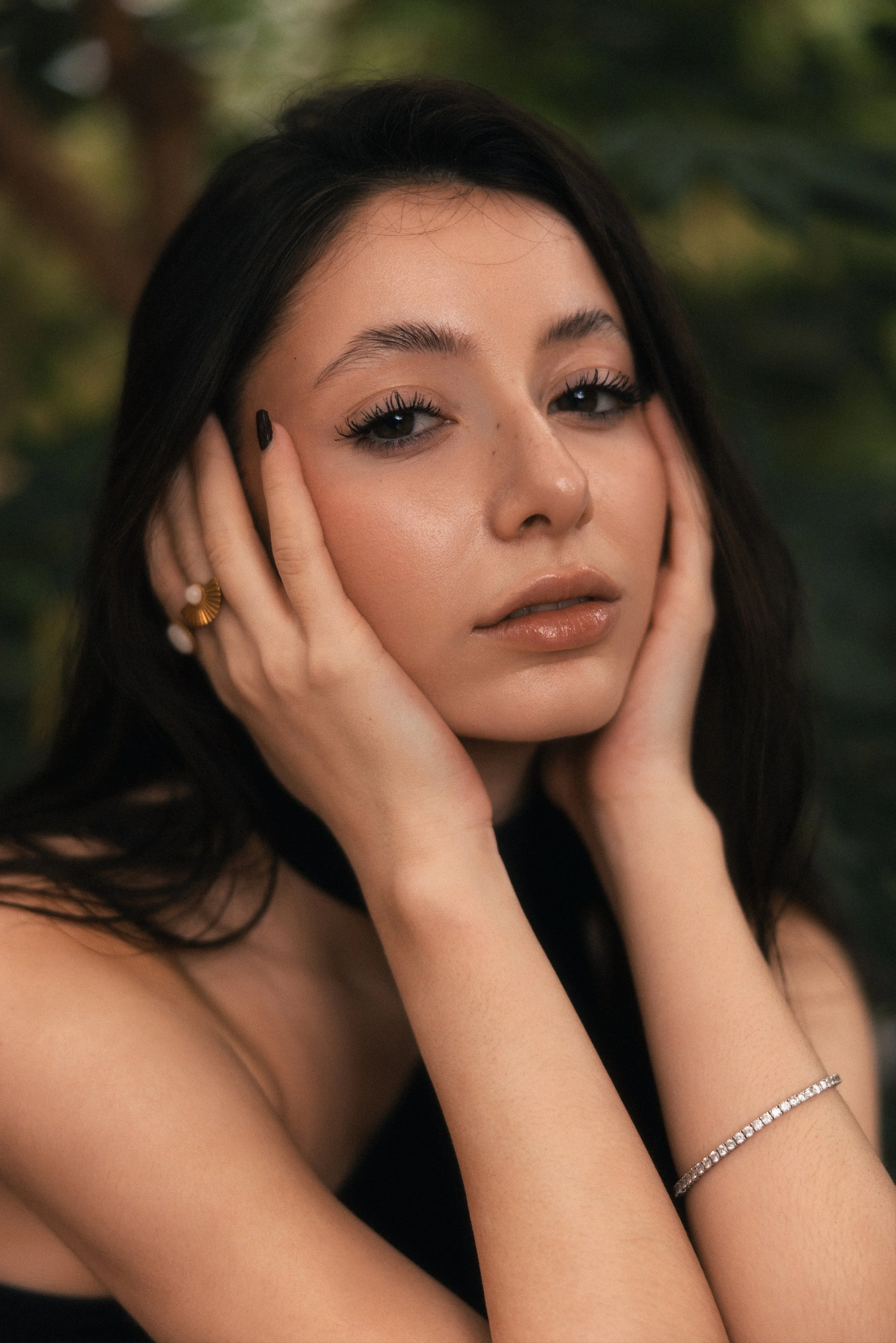 Close-up of a young woman with dark hair, light makeup, and jewelry, resting her face in her hands with a blurred green background.