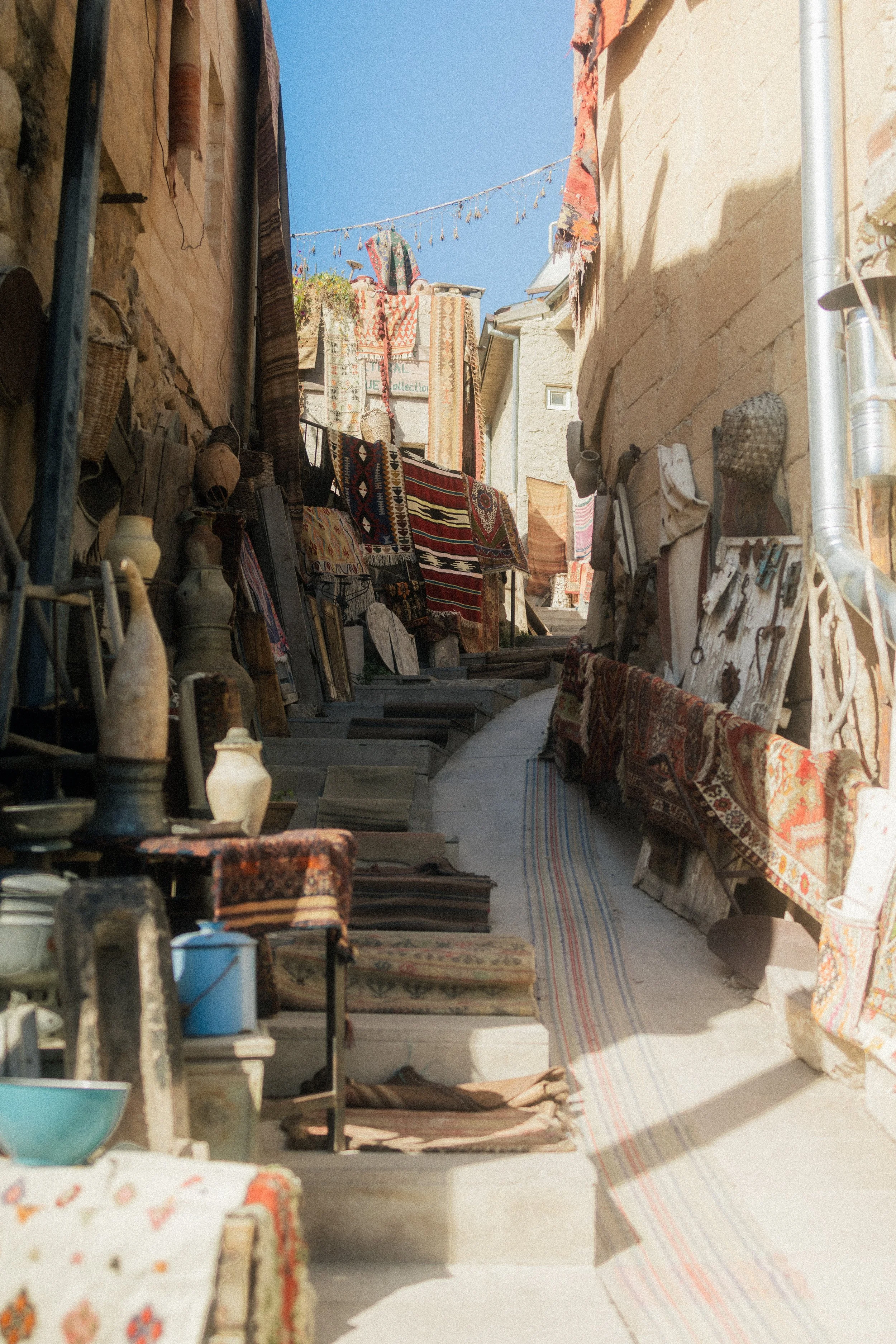 An alleyway with stone walls on both sides, decorated with hanging carpets, rugs, and textiles, with a clear blue sky overhead.