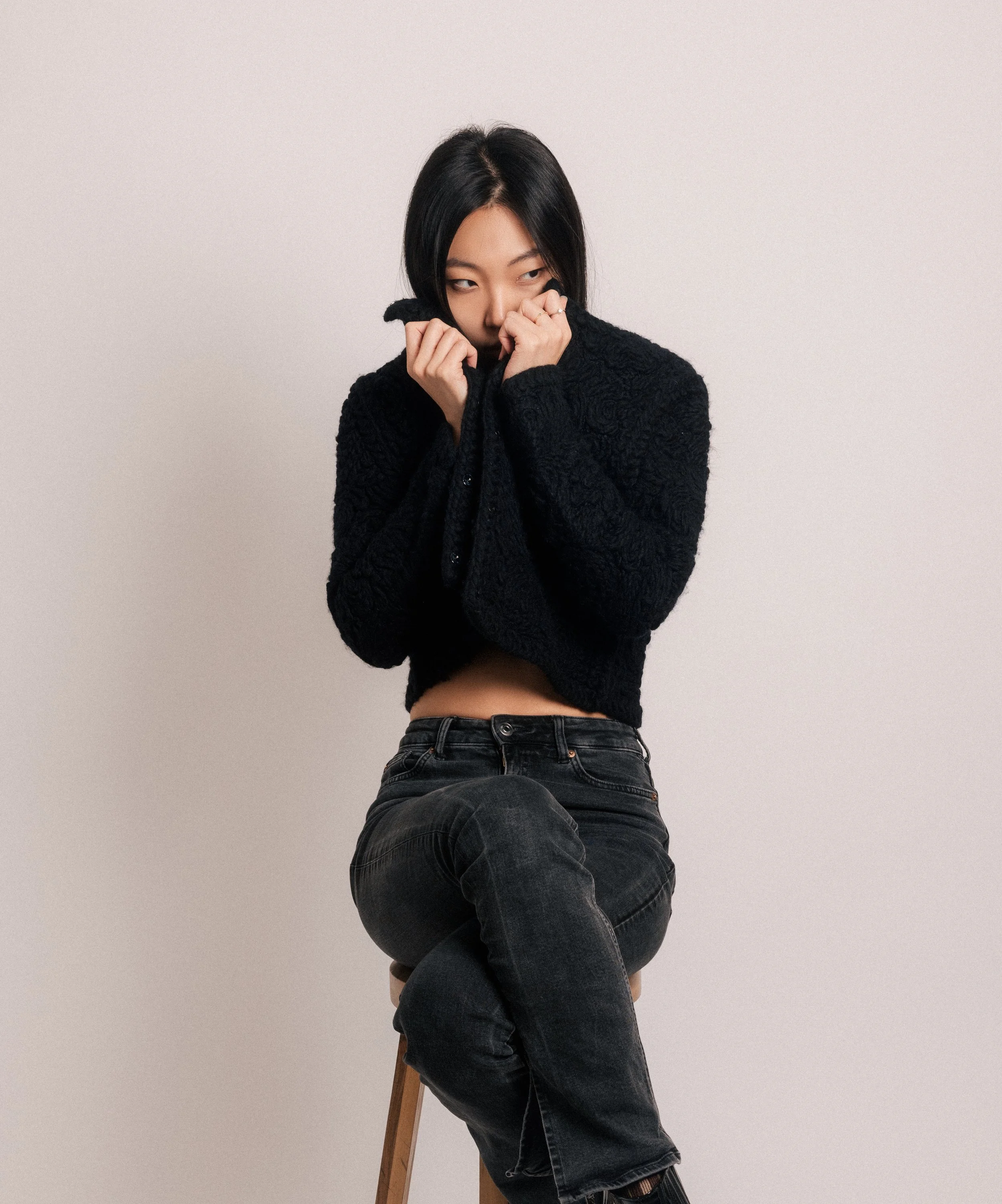 A woman with black hair wearing a black sweater and jeans is sitting on a stool, holding the collar of her sweater up to her nose against a plain, light-colored background.