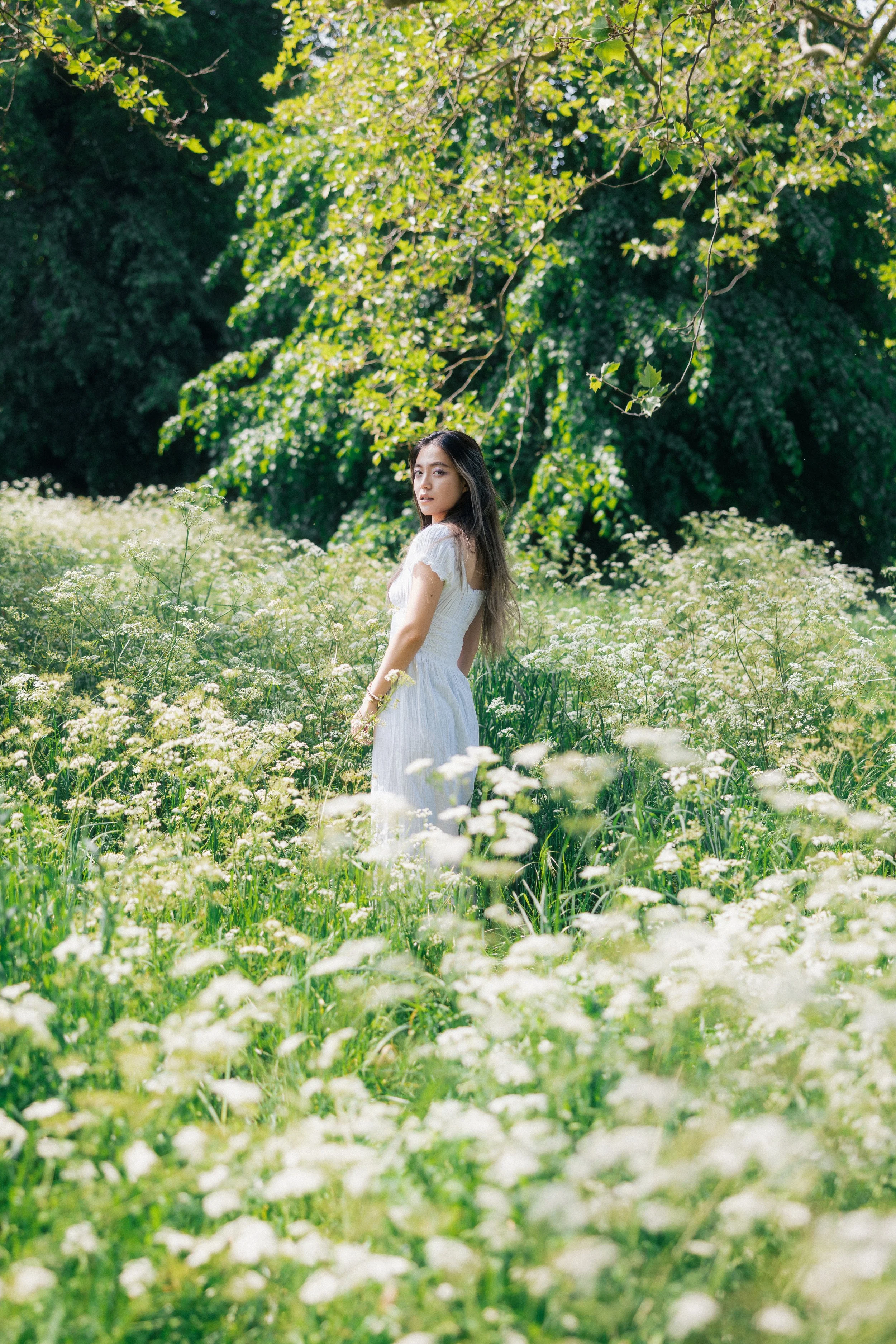 A young woman in a white dress standing in a field of white flowers surrounded by green trees.