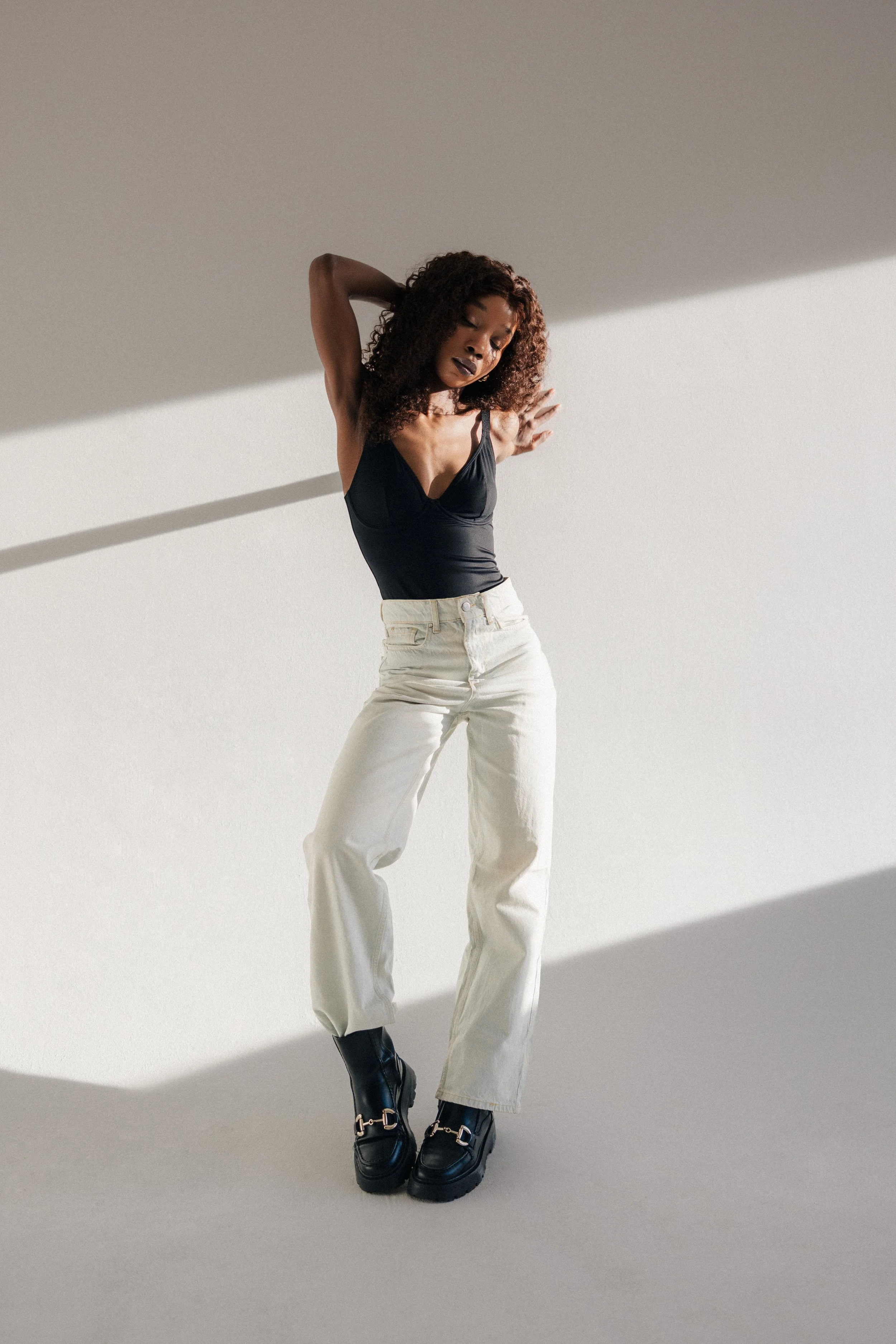A woman with curly hair standing with one arm raised behind her head and the other crossed over her shoulder, wearing a black tank top, white pants, and black boots with metal buckle details, in a minimalist setting with shadows cast on the wall.