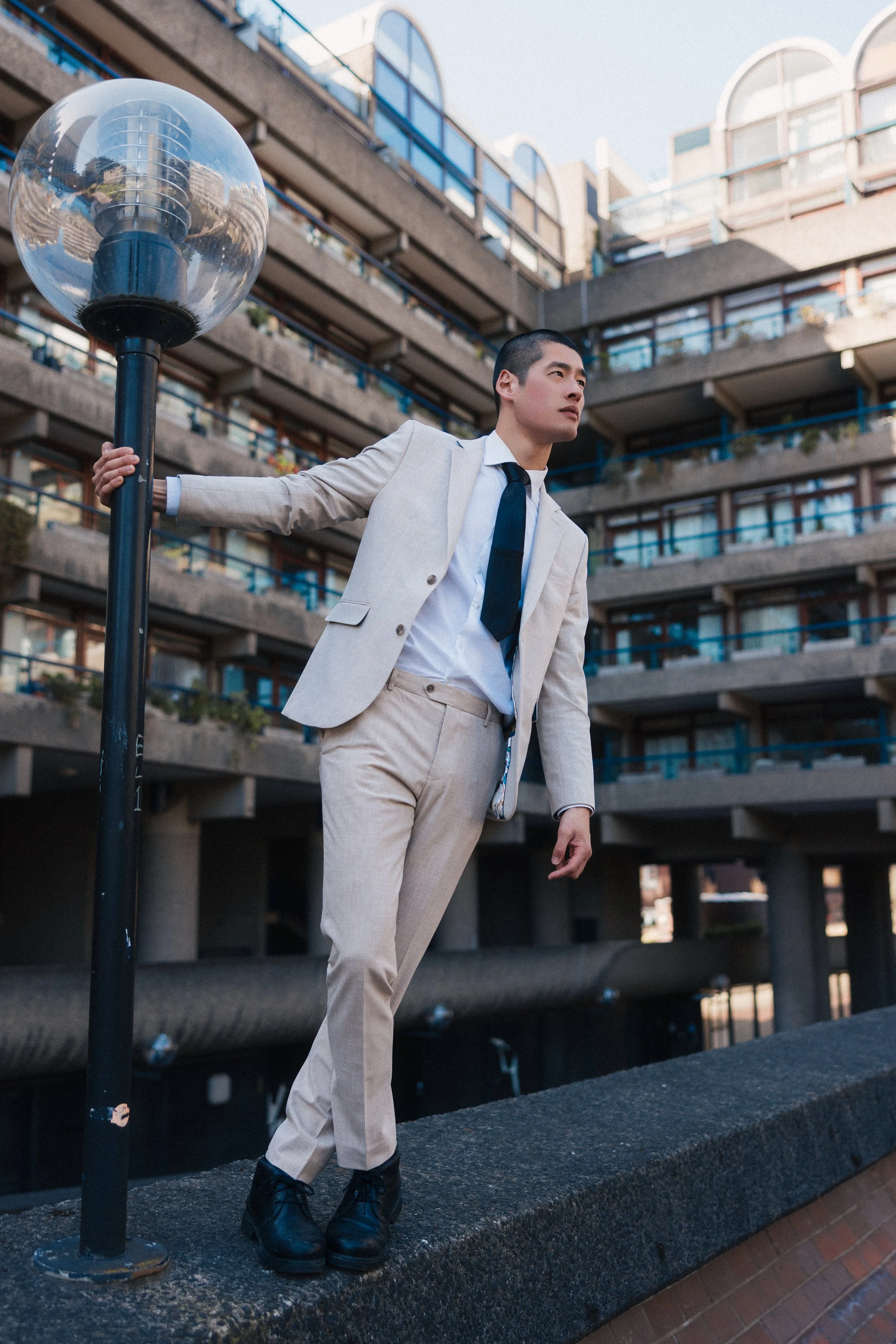A man dressed in a light-colored suit, white dress shirt, and dark tie, wearing black boots, standing outdoors next to a modern urban building with multiple balconies, holding onto a streetlamp post, looking into the distance.