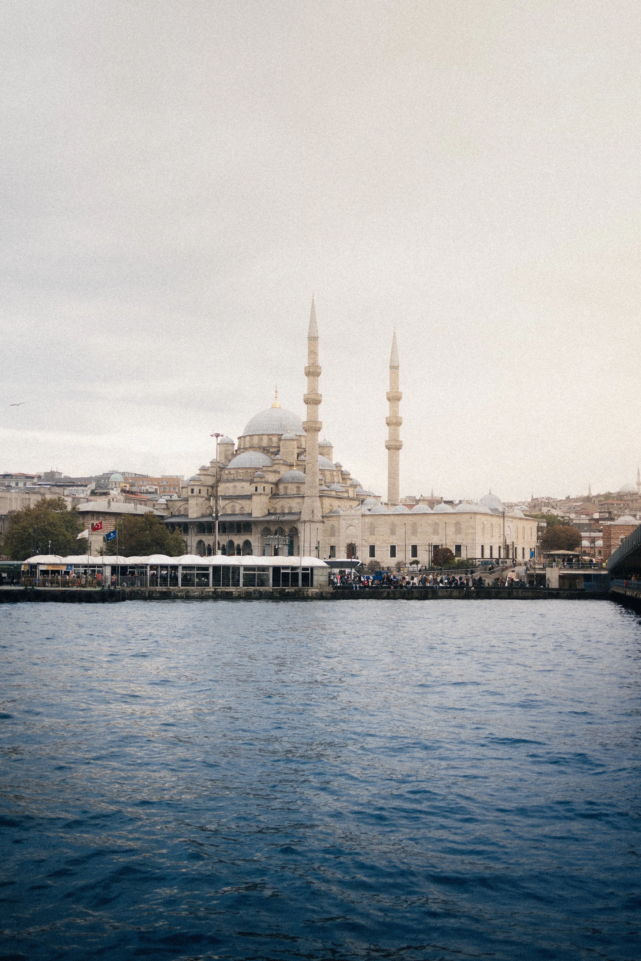 View of a mosque with minarets and domes along a waterway, under an overcast sky in an urban area.