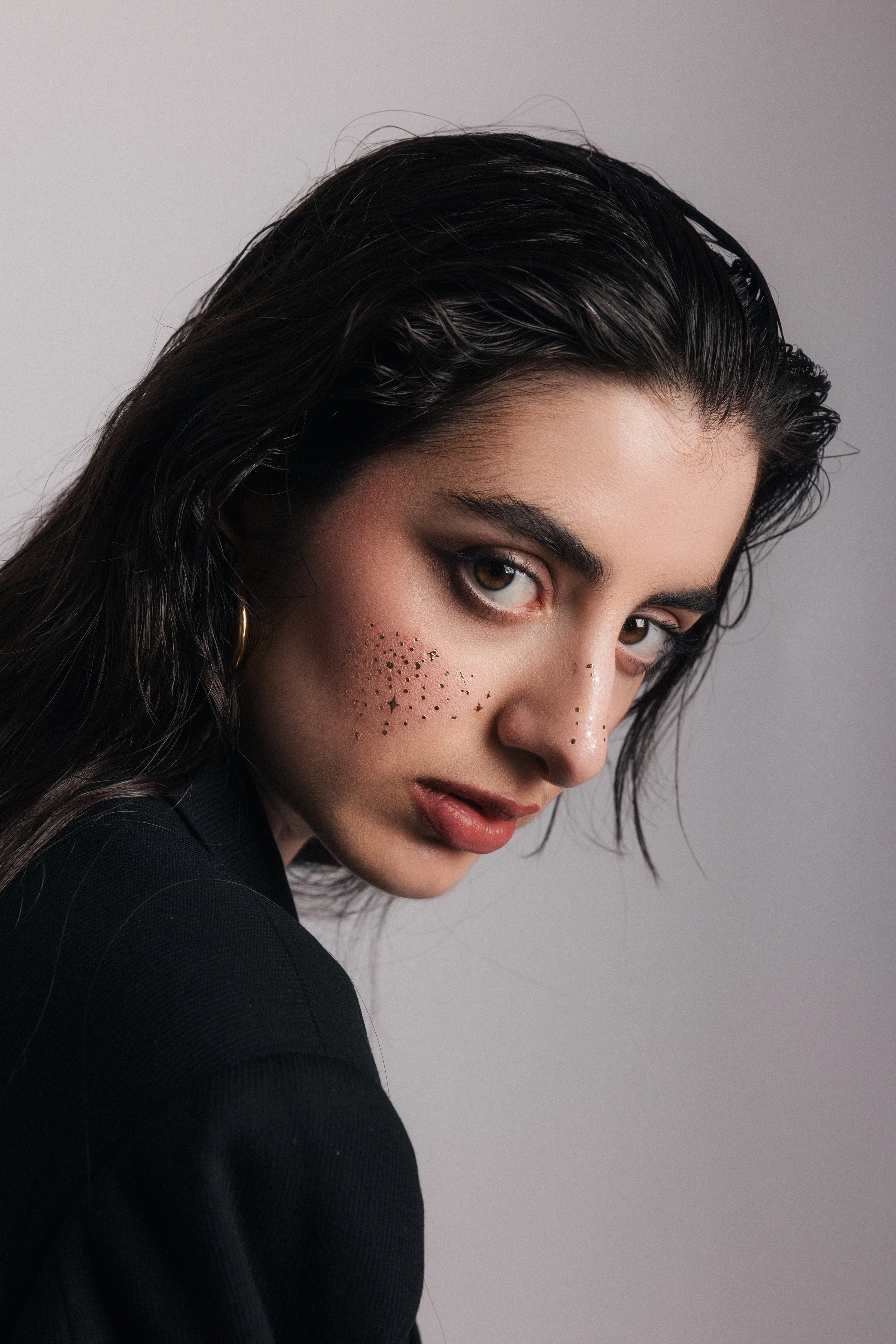 Close-up of a woman with dark wet hair, light brown eyes, natural makeup, and gold hoop earrings, wearing a black top against a gray background.