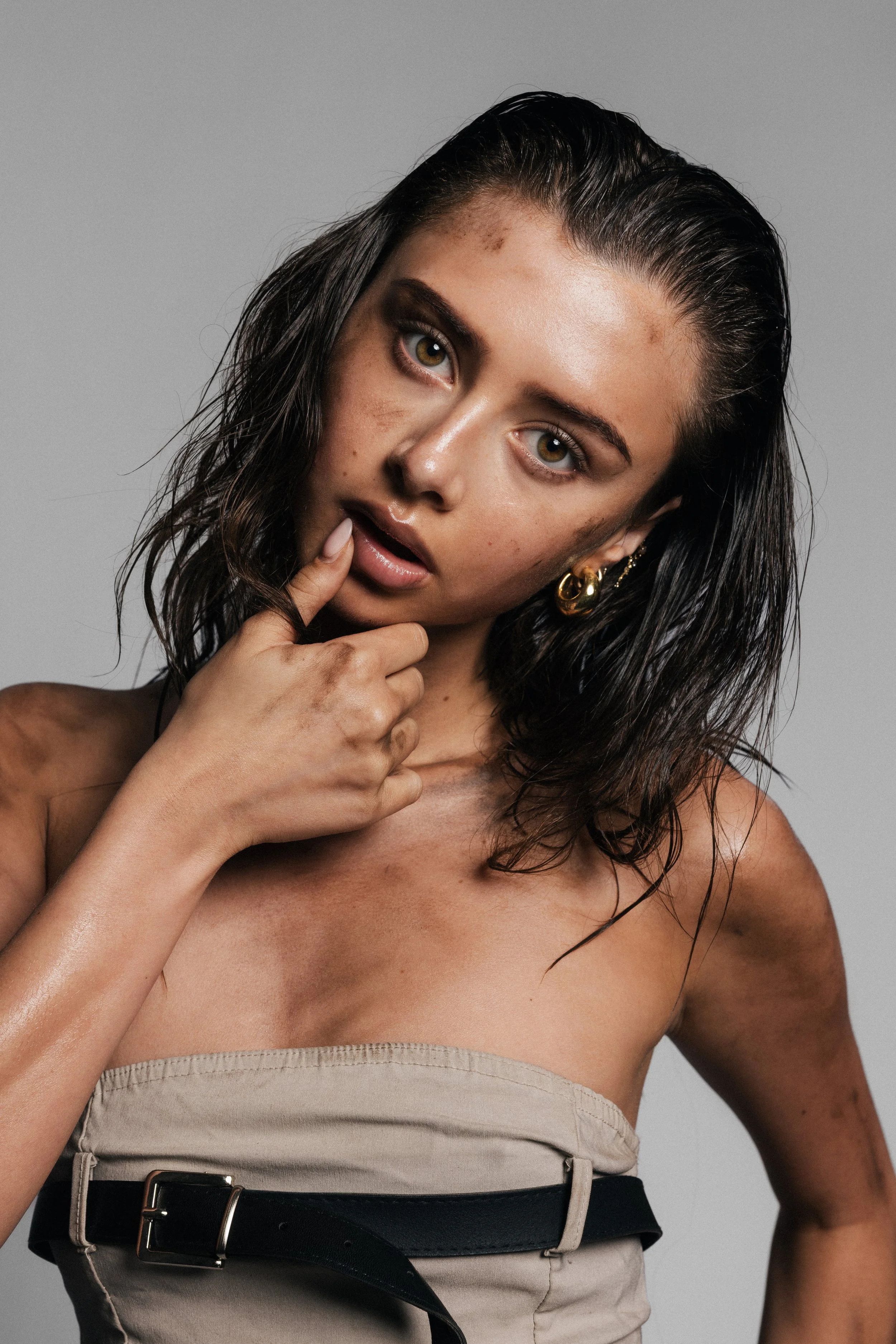 A woman with wet black hair, light brown eyes, and tan skin, wearing a beige strapless top and a black belt with metal buckle, posing against a plain gray background, touching her chin with her left hand.