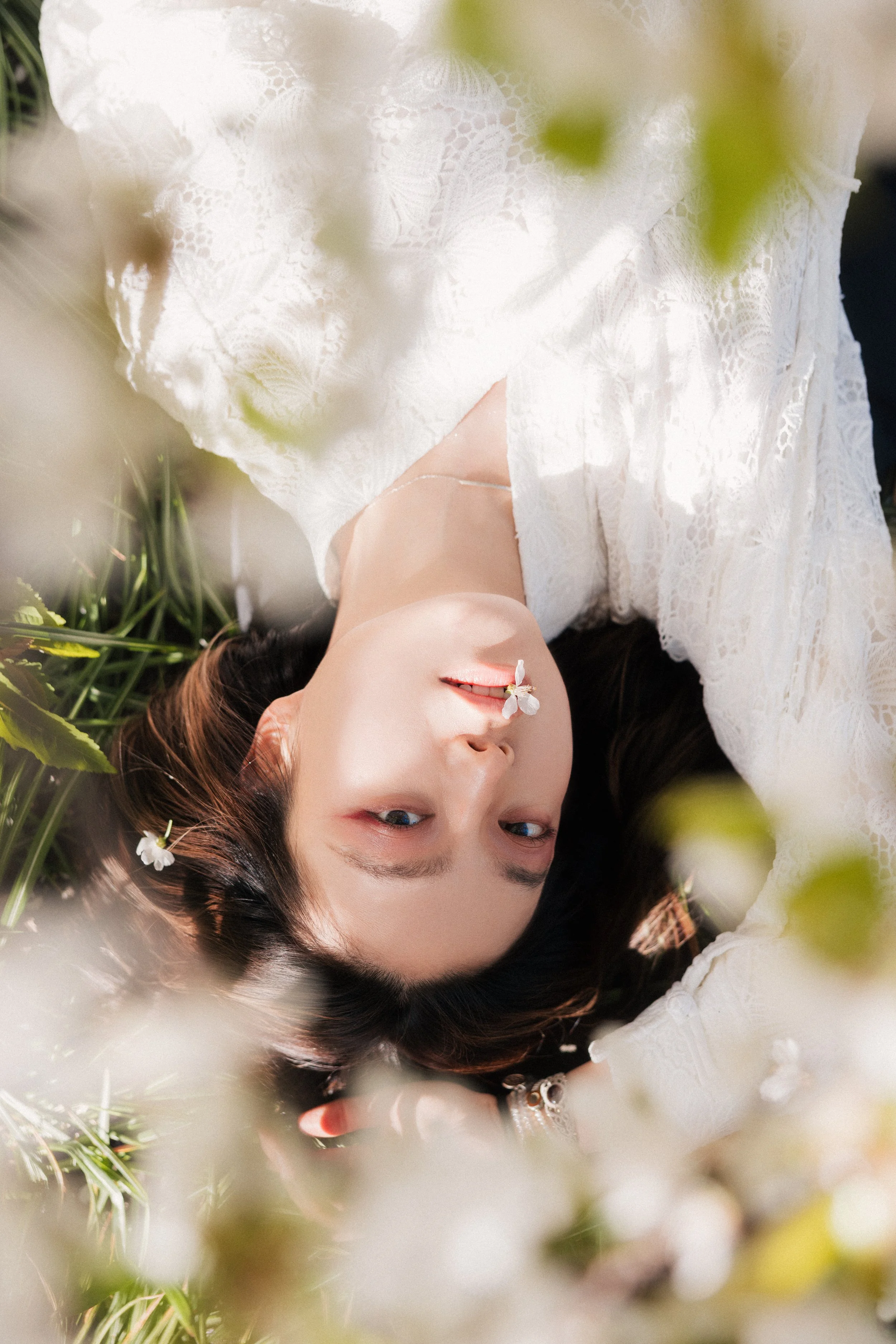 A woman lying on grass, surrounded by white flowers, wearing a white lace dress, with a small white flower on her face.