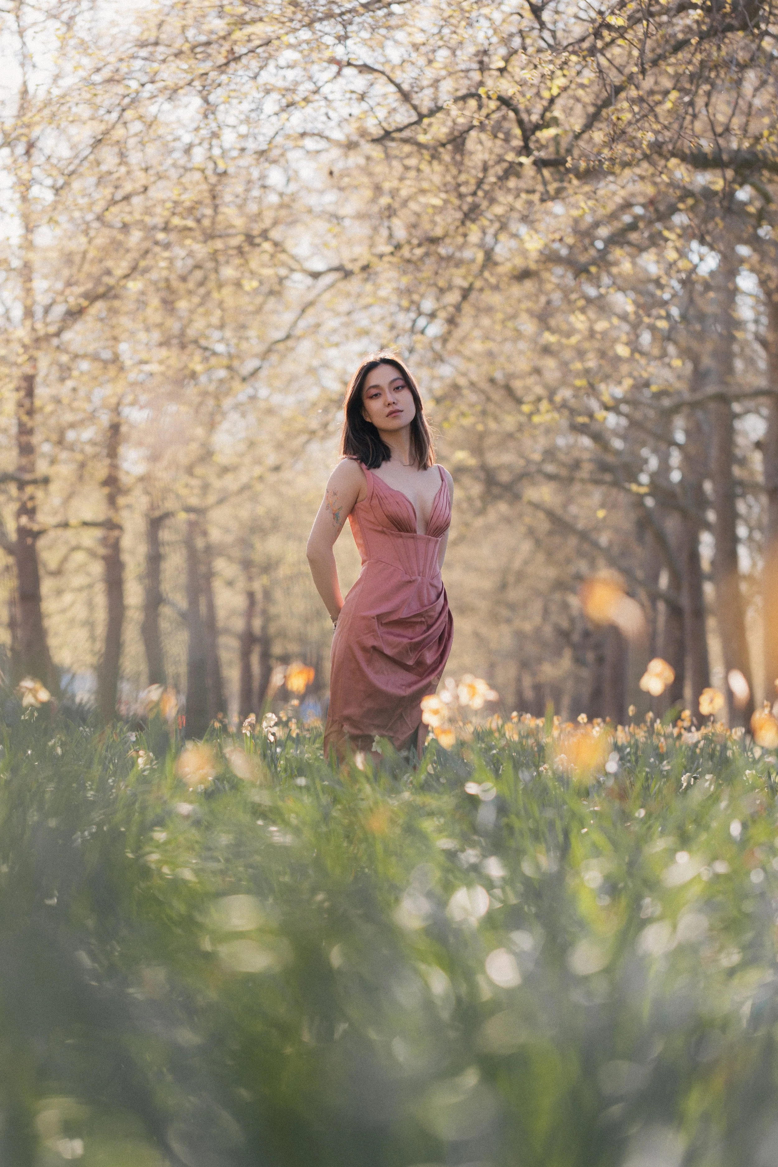 A woman standing in a field of yellow flowers with a backdrop of trees with yellow autumn leaves, wearing a pink dress