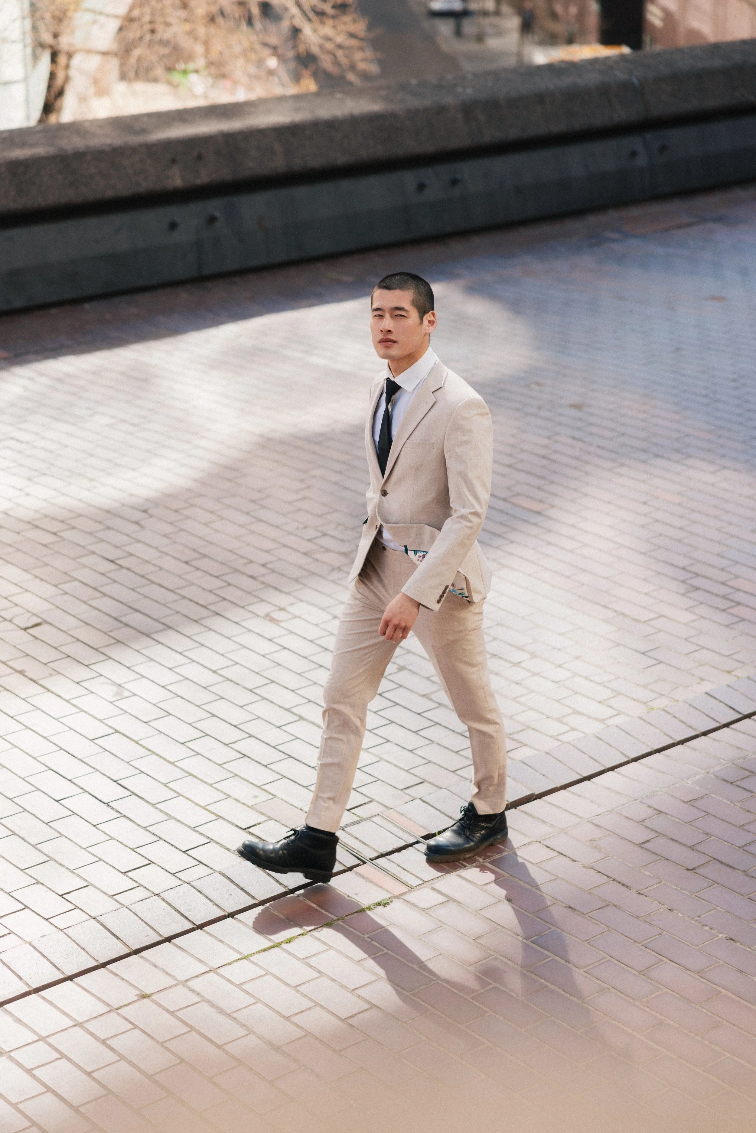 A man in a beige suit with a white shirt and black tie walking on a paved sidewalk, wearing black shoes and looking at the camera.