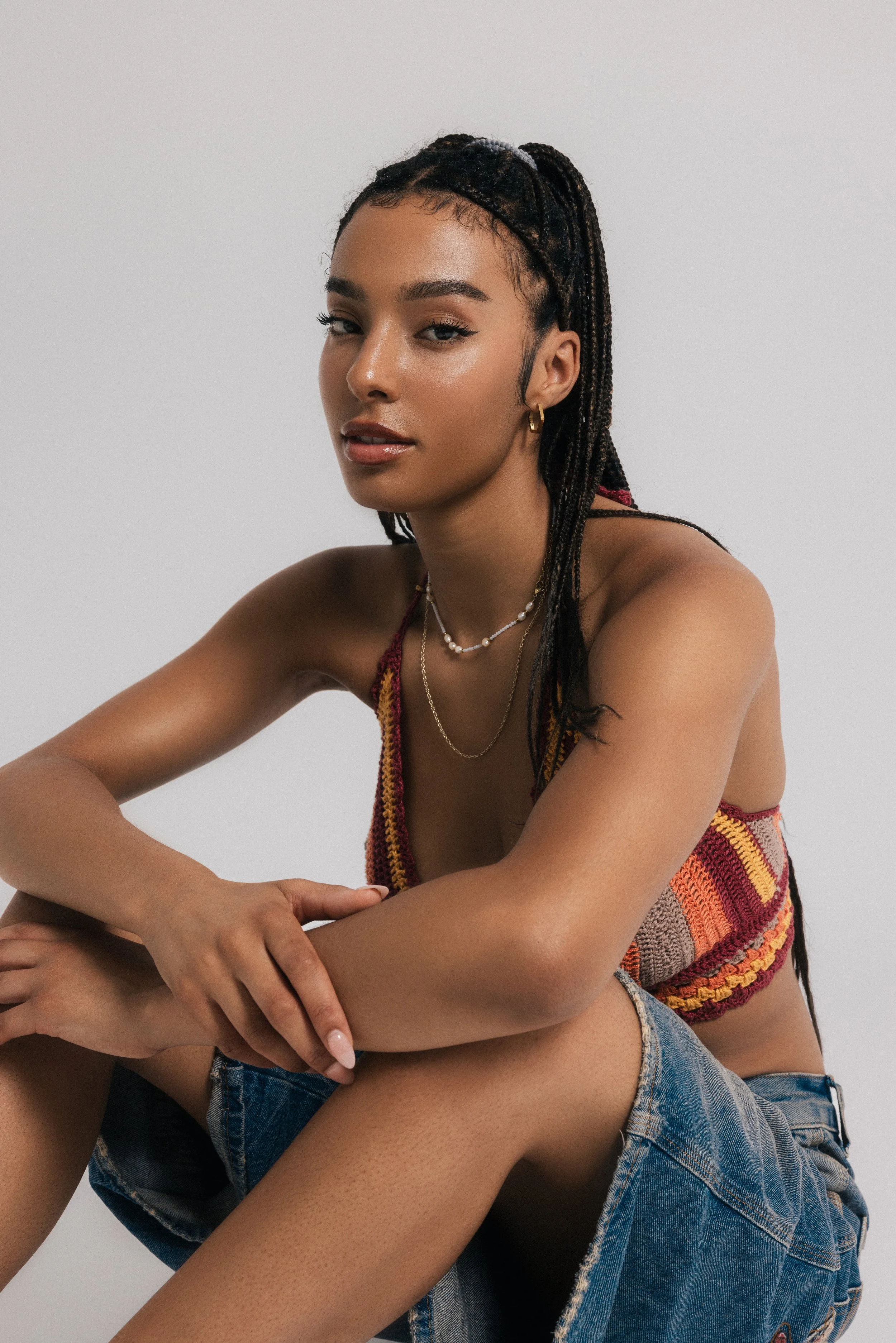 Portrait of a young woman with braided hair, wearing jewelry and a colorful knitted top, against a plain white background.