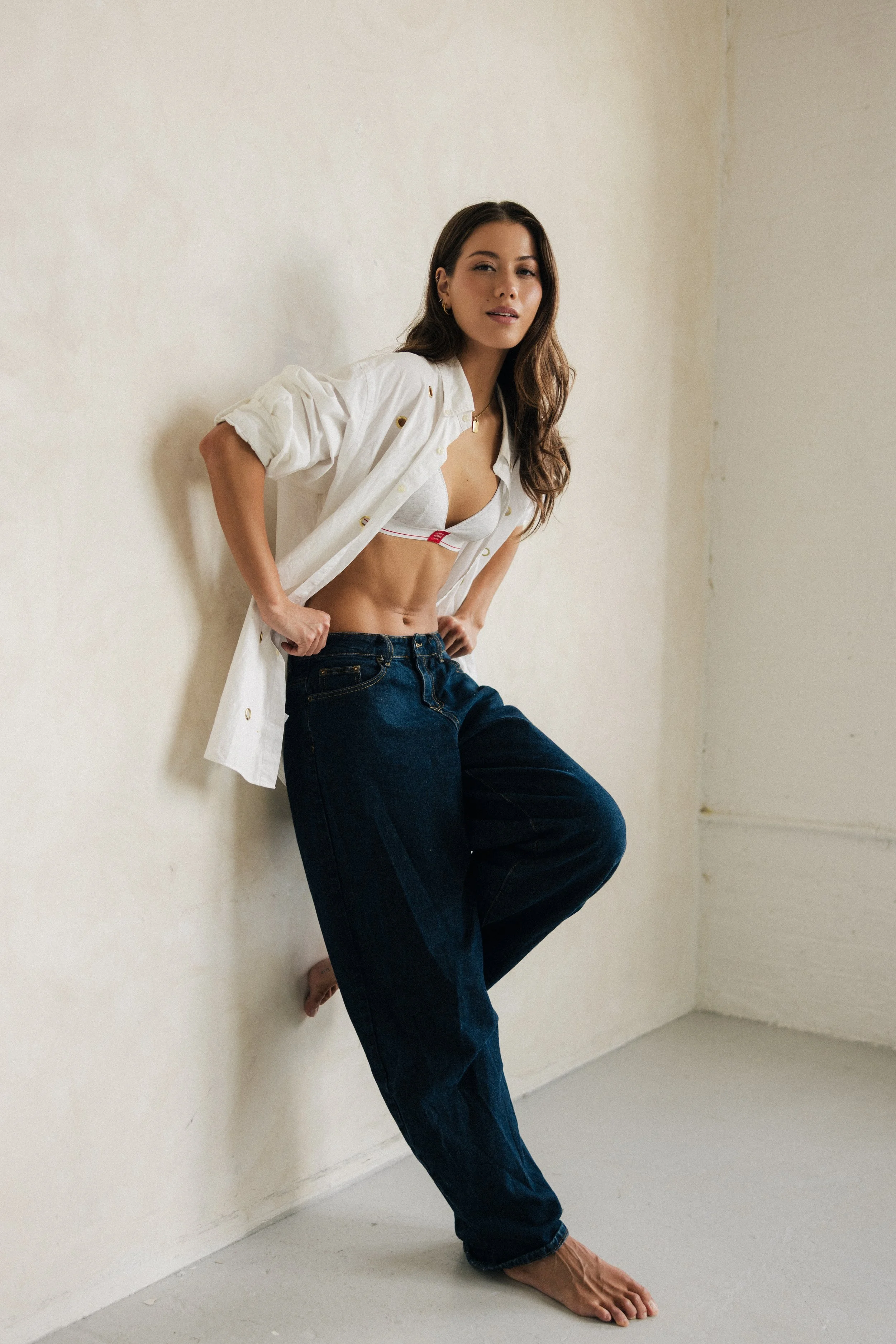 A woman with long brown hair and light skin leaning against a plain off-white wall. She is wearing a white open shirt over a white sports bra, dark blue jeans, and is barefoot. Her right hand rests on her hip, and her left hand is placed on her jeans