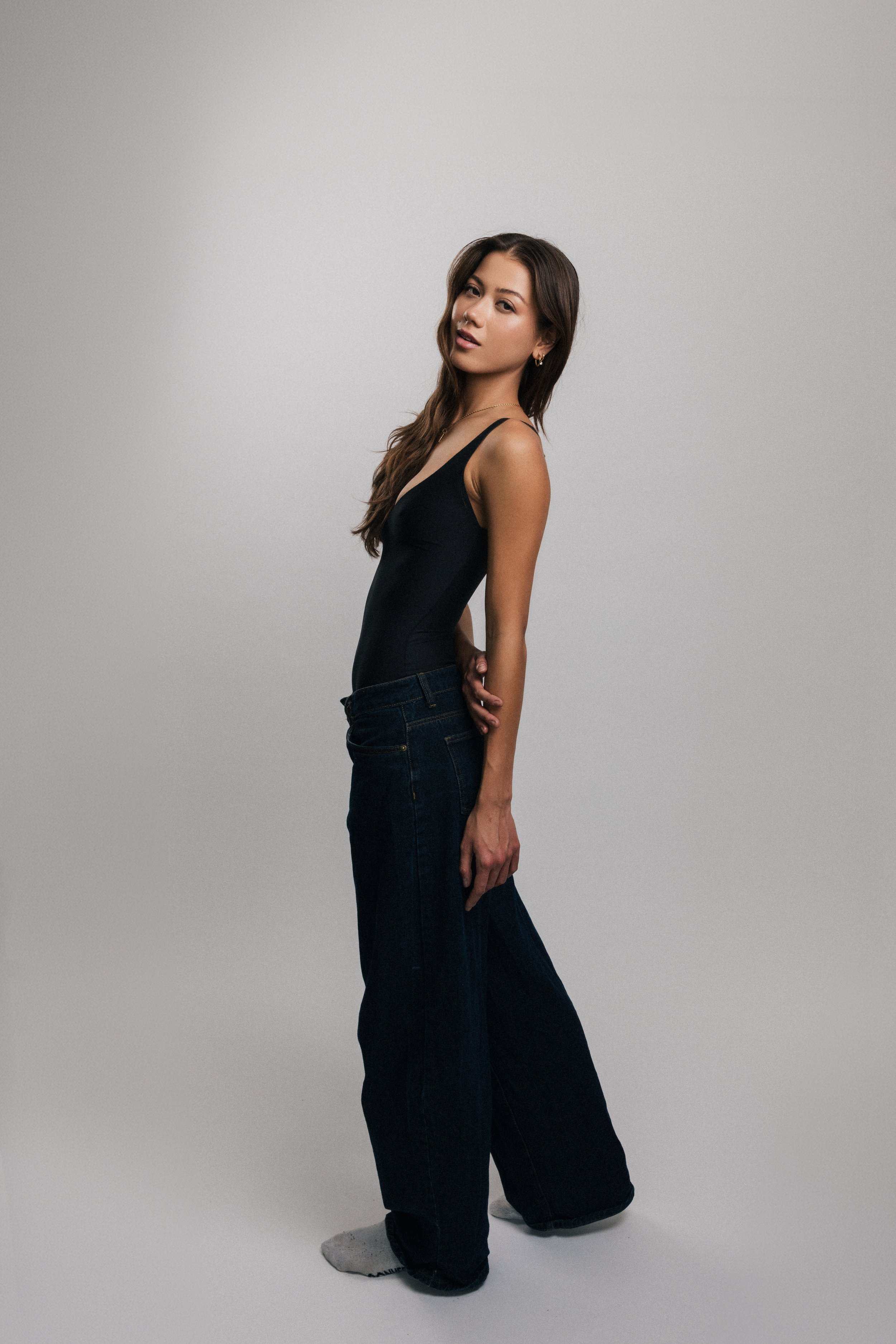 Young woman with long brown hair wearing black tank top and dark jeans standing against a plain gray background.