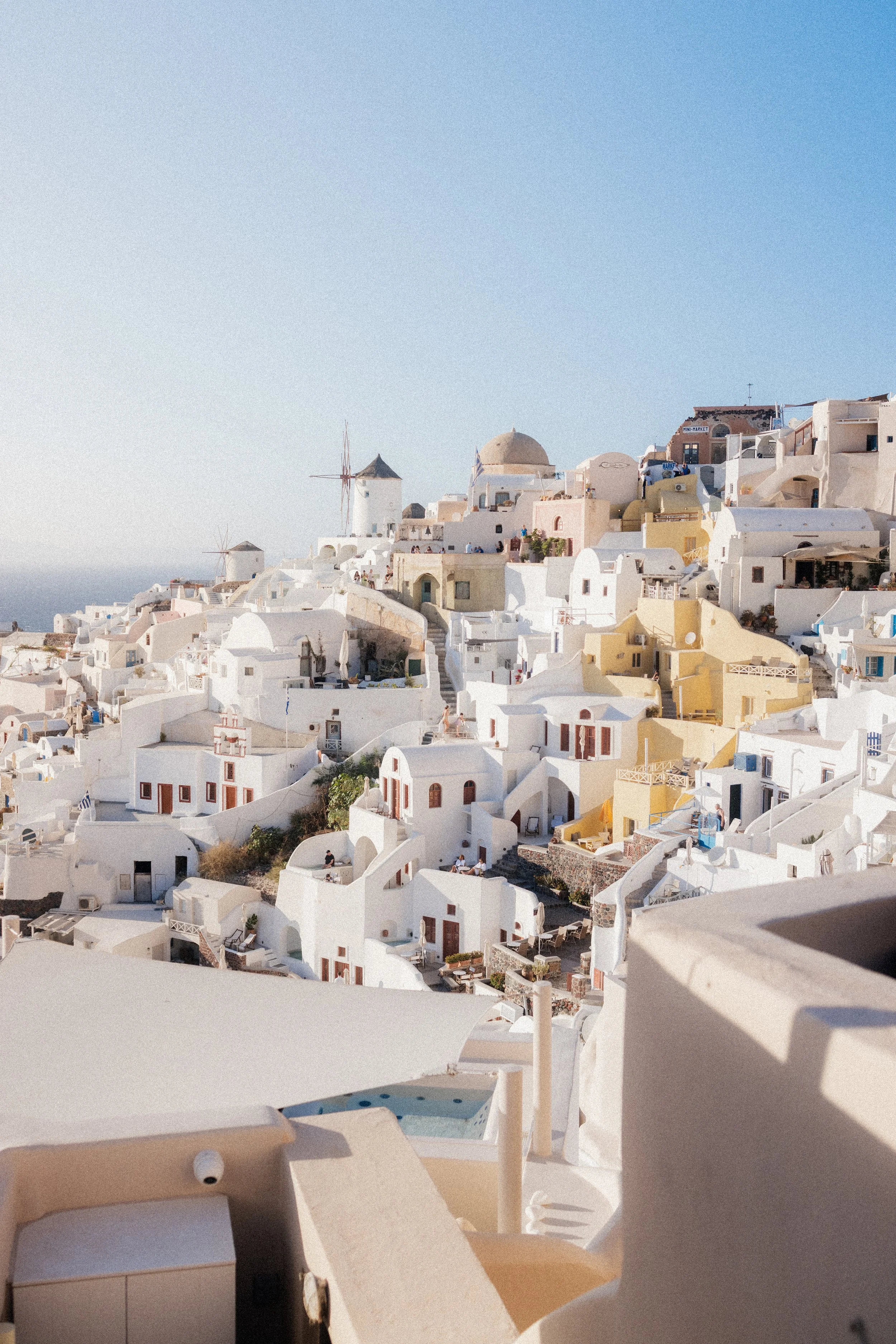 White buildings with rounded shapes and small windows in a coastal town, with windmills and a clear blue sky in the background, characteristic of Santorini, Greece.