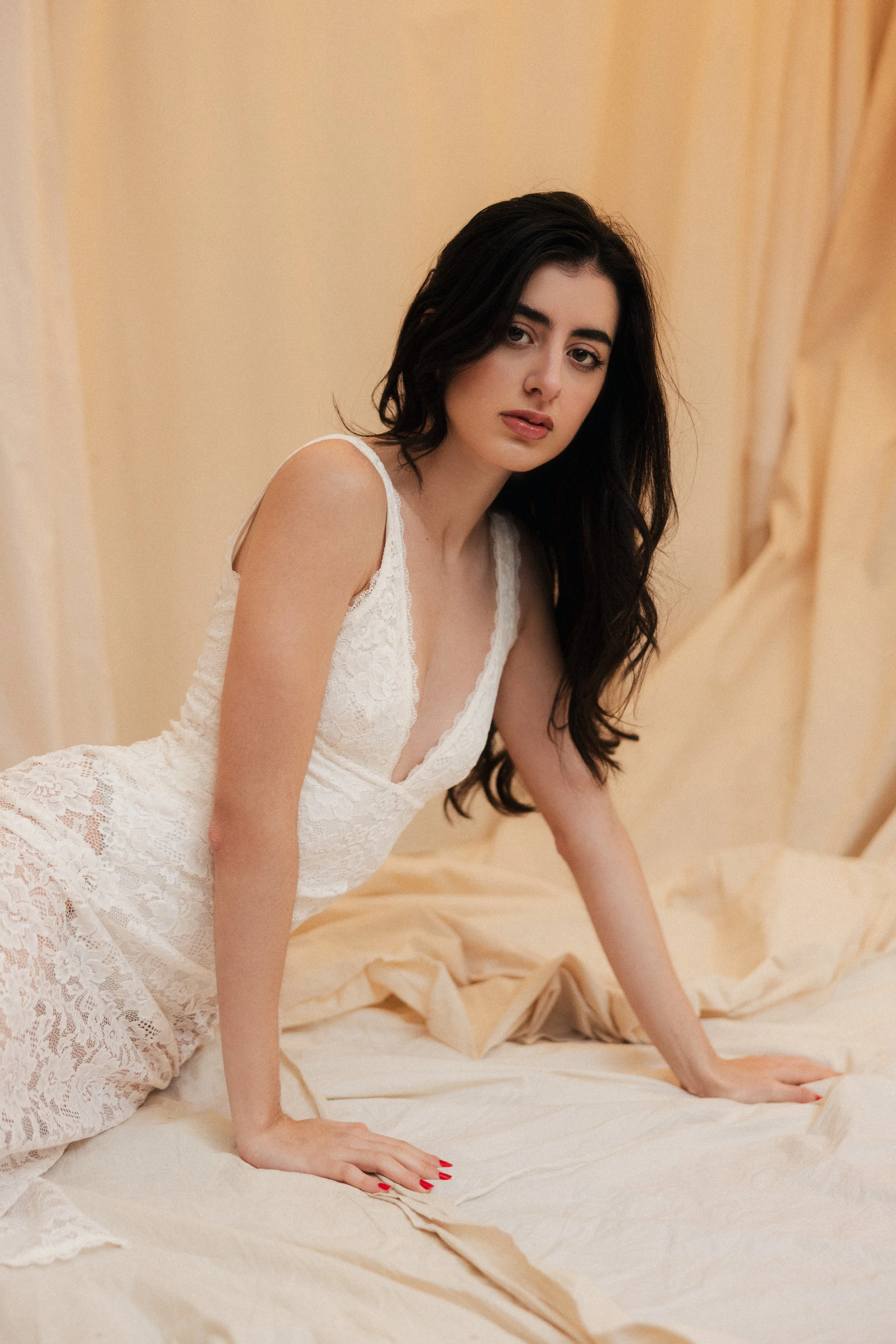 A woman with long dark hair in a white lace dress is on her hands and knees on a bed with beige sheets, looking at the camera.