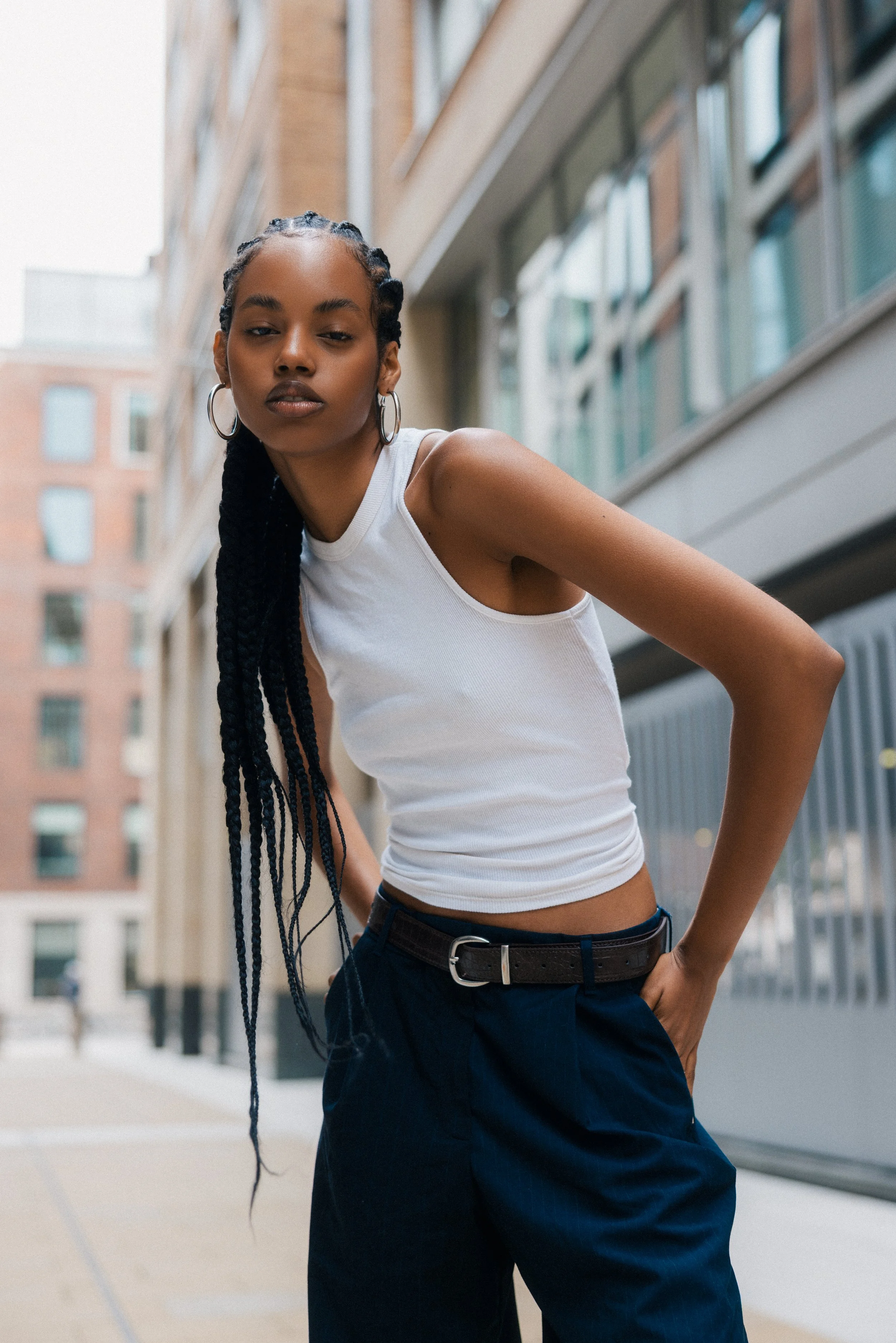 A young woman with long braided hair wearing hoop earrings, a white tank top, and dark pants standing outdoors in an urban area with modern buildings in the background.