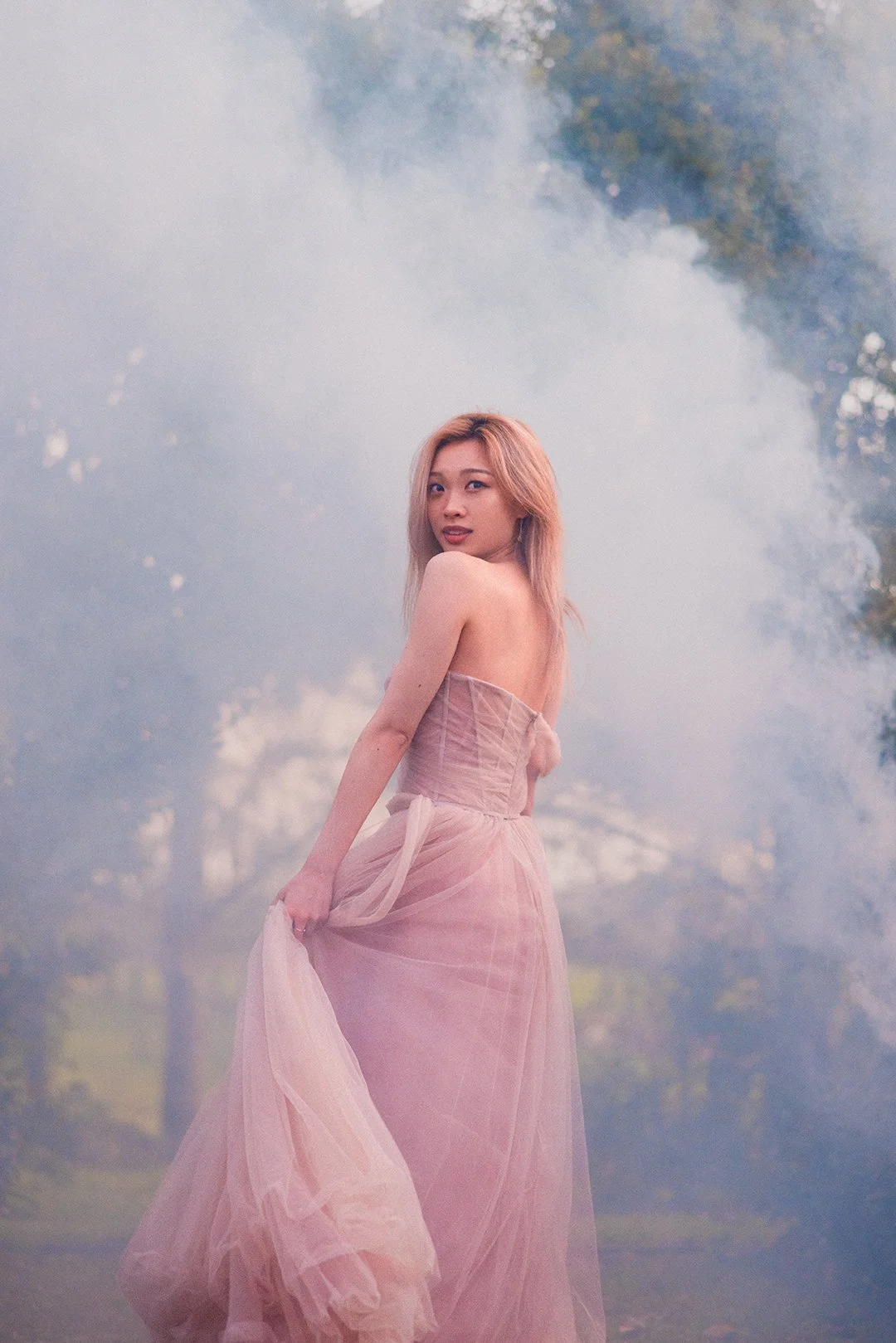 A woman in a flowing pink dress standing in a foggy outdoor setting with trees in the background.