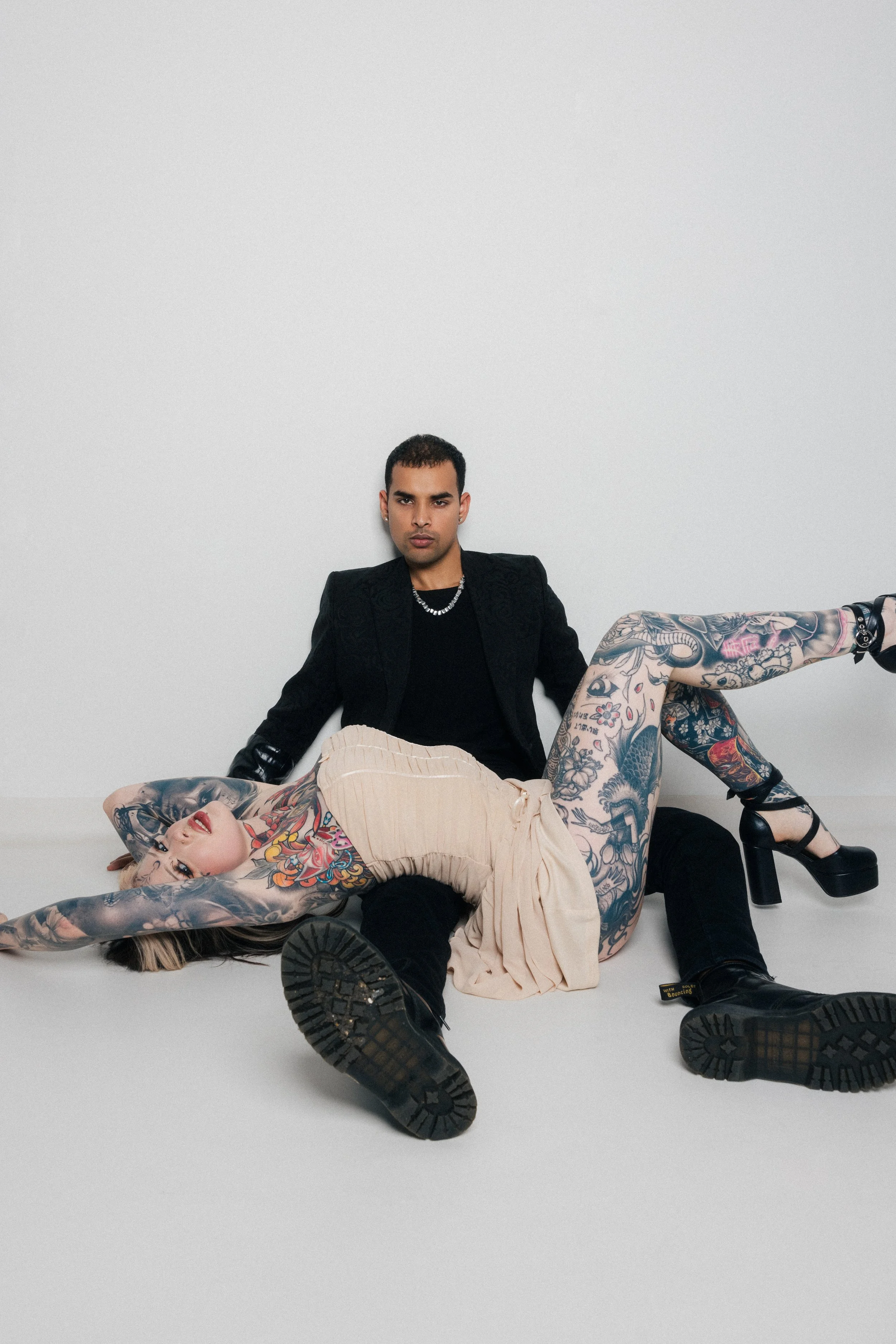 Two individuals with extensive tattoos, one lying on the floor and the other sitting against a plain white wall. The person on the floor is a woman with dark makeup, long hair, and colorful tattoos, wearing a beige dress. The person sitting is a man 