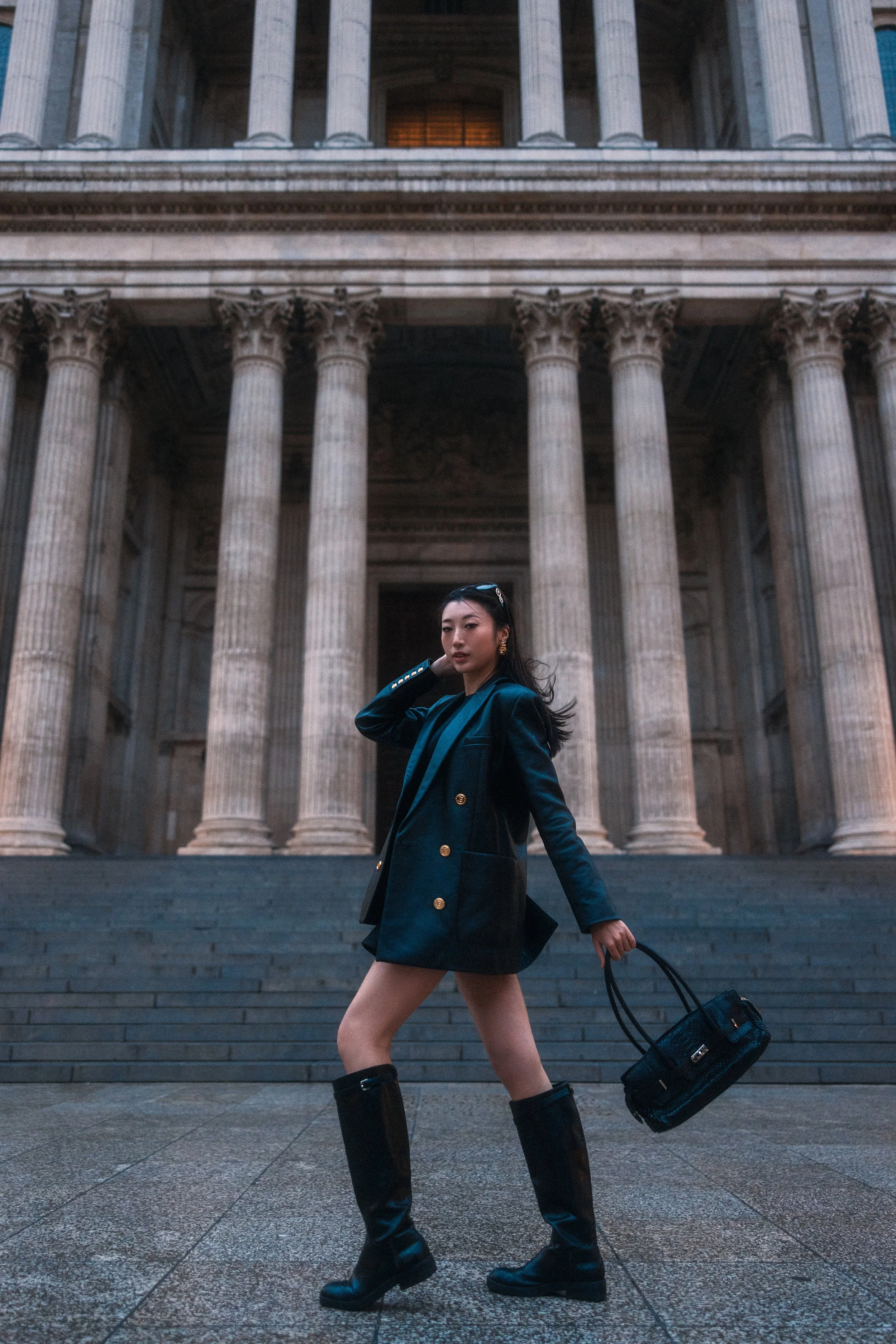 A woman in a dark teal blazer and black knee-high boots walking down steps in front of a building with classical architecture, large columns.