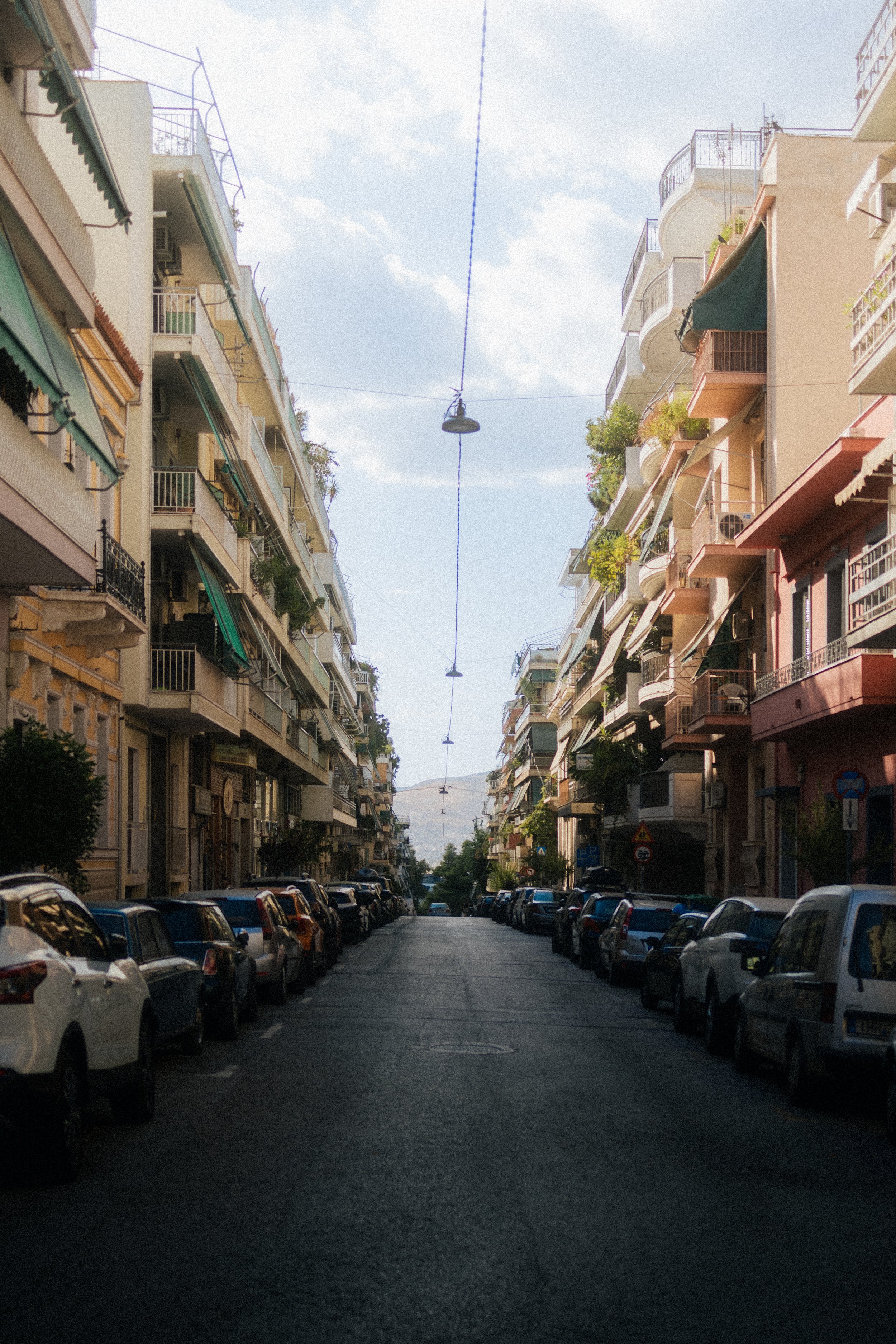 View of a narrow city street flanked by multi-story apartment buildings with balconies and awnings. Cars are parked on both sides of the street, and a streetlight hangs overhead against a partly cloudy sky.