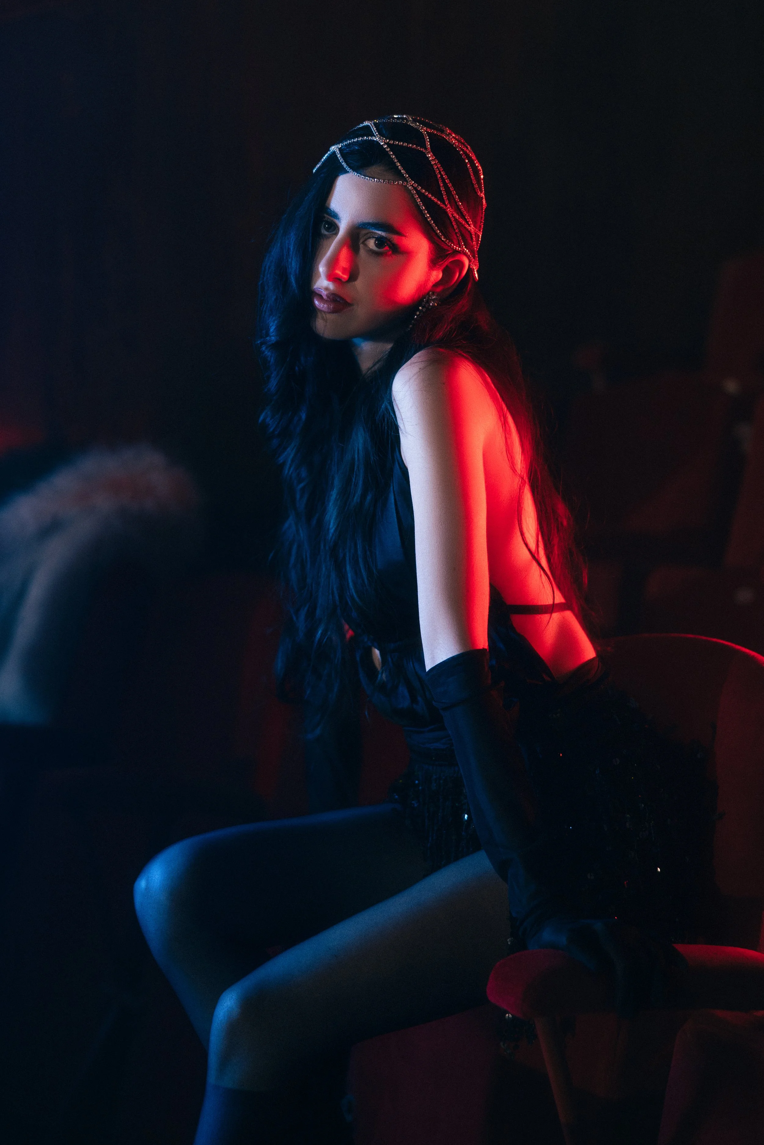 A woman with long black hair, wearing a beaded headpiece, black dress, and black gloves, sitting in a dark setting illuminated with red and blue lighting, looking at the camera.