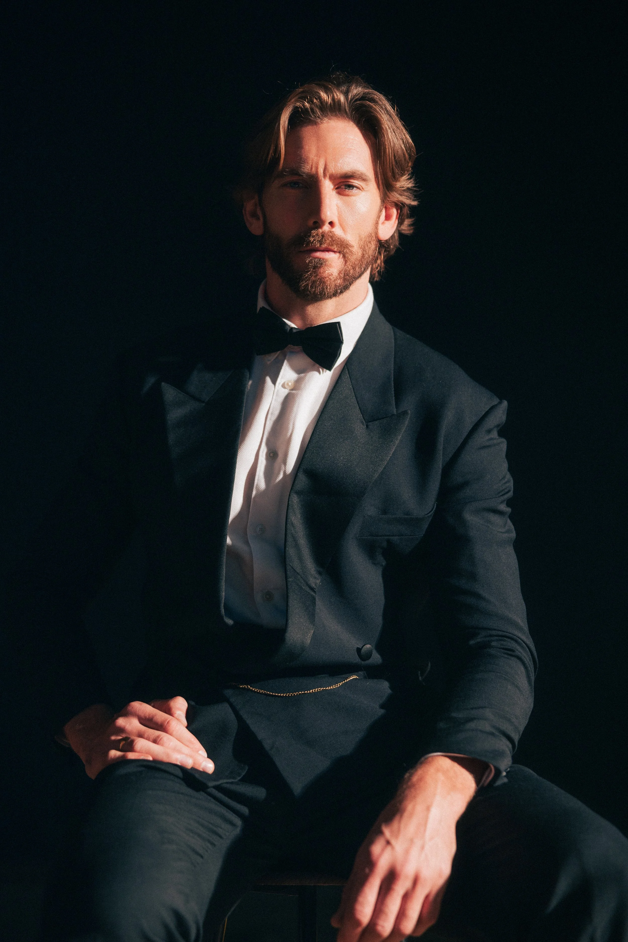 A man with light skin, a beard, and long hair styled to one side, wearing a black tuxedo with a white shirt and a black bowtie, sits against a dark background.