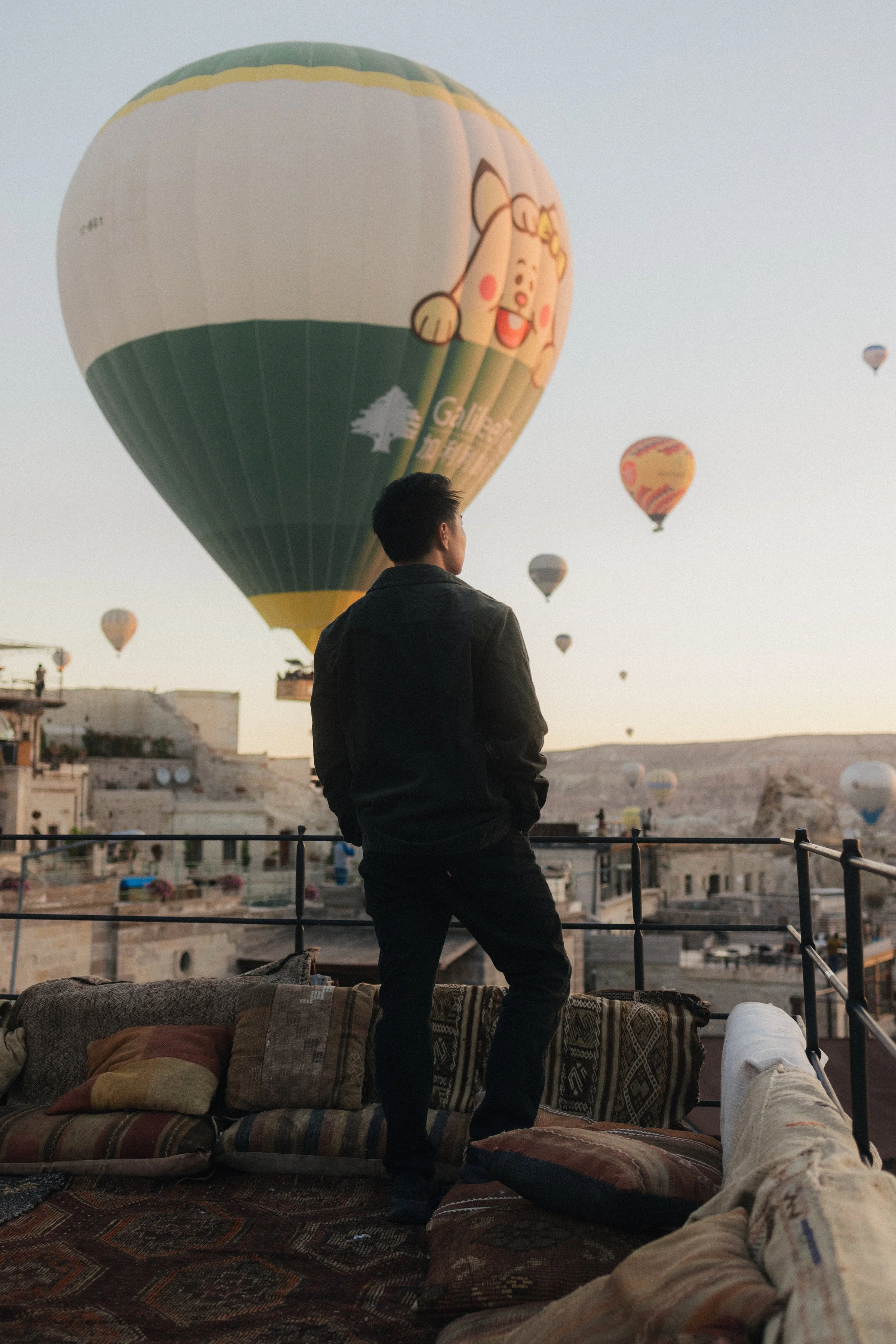 A man in a dark jacket and pants standing on a rooftop balcony, looking at hot air balloons in the sky during sunset, with cushions and a person lying on the ground in front of him.
