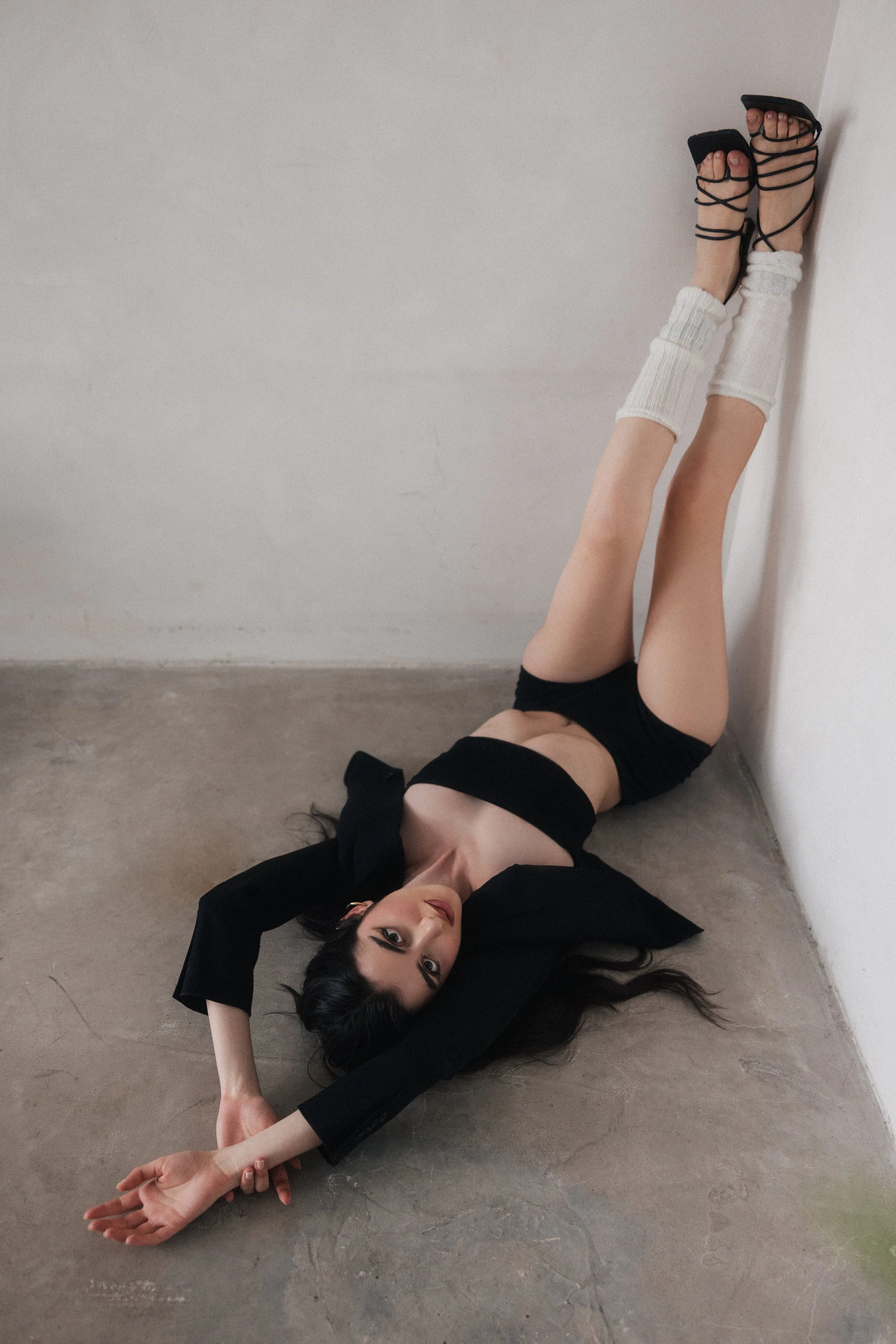 A woman lying on the floor against a white wall, wearing black clothing, white knee-high socks, and black high-heeled sandals with straps. She has black hair and is looking at the camera.