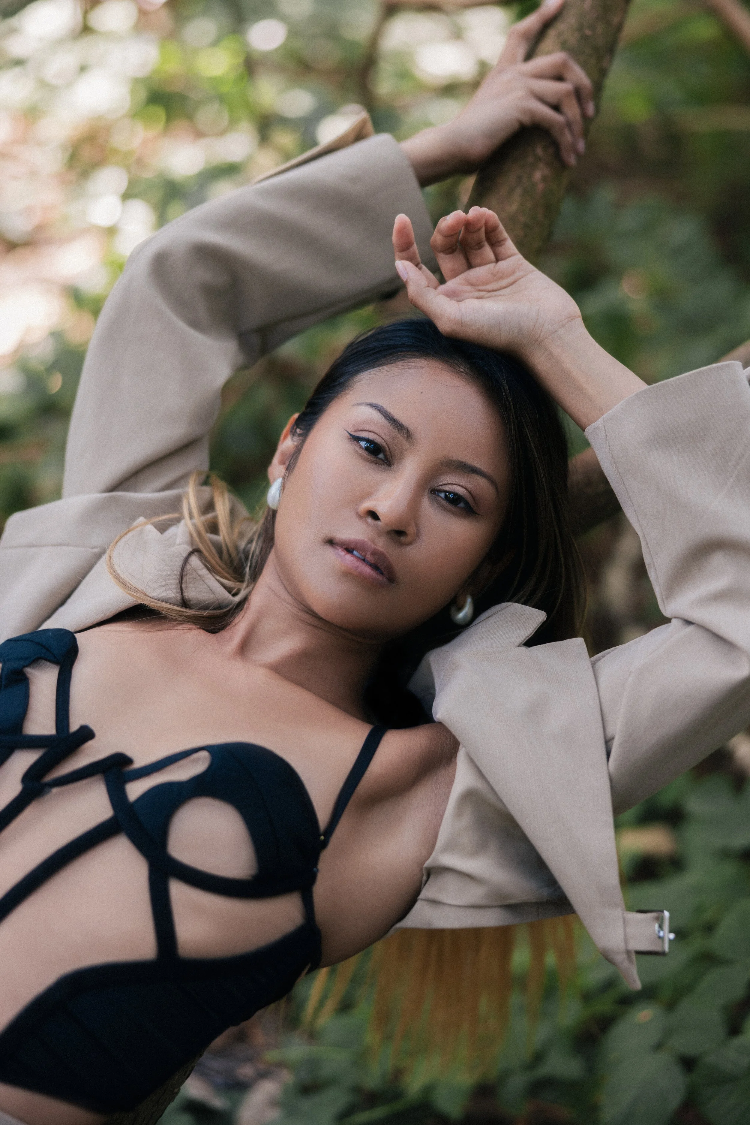 A woman lying outdoors next to a tree, with her arm resting on the tree branch and her other hand touching her forehead. She is wearing a beige coat and a black lace-up body suit, with earrings and makeup.