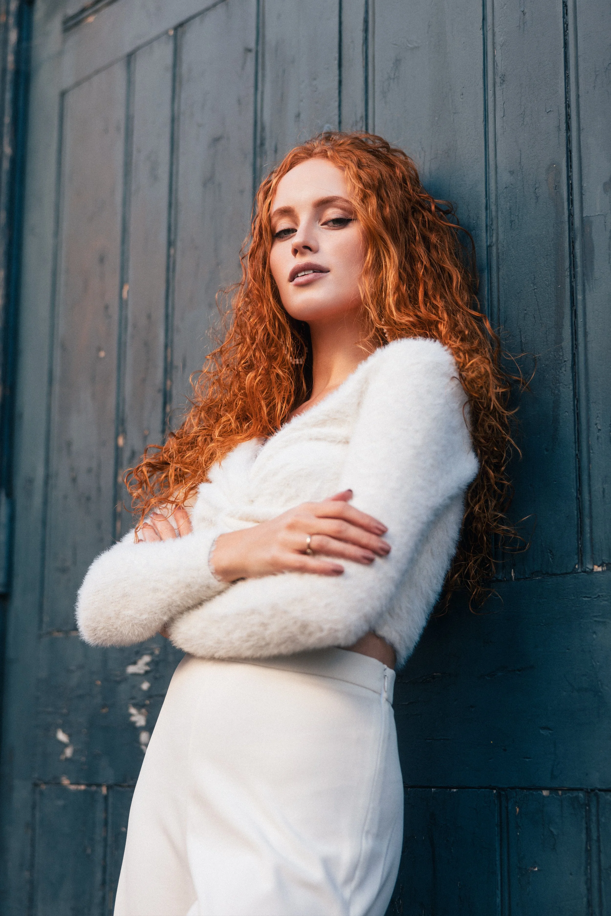 A woman with long curly red hair wearing a white fuzzy sweater and white pants, standing against a dark blue wooden wall.