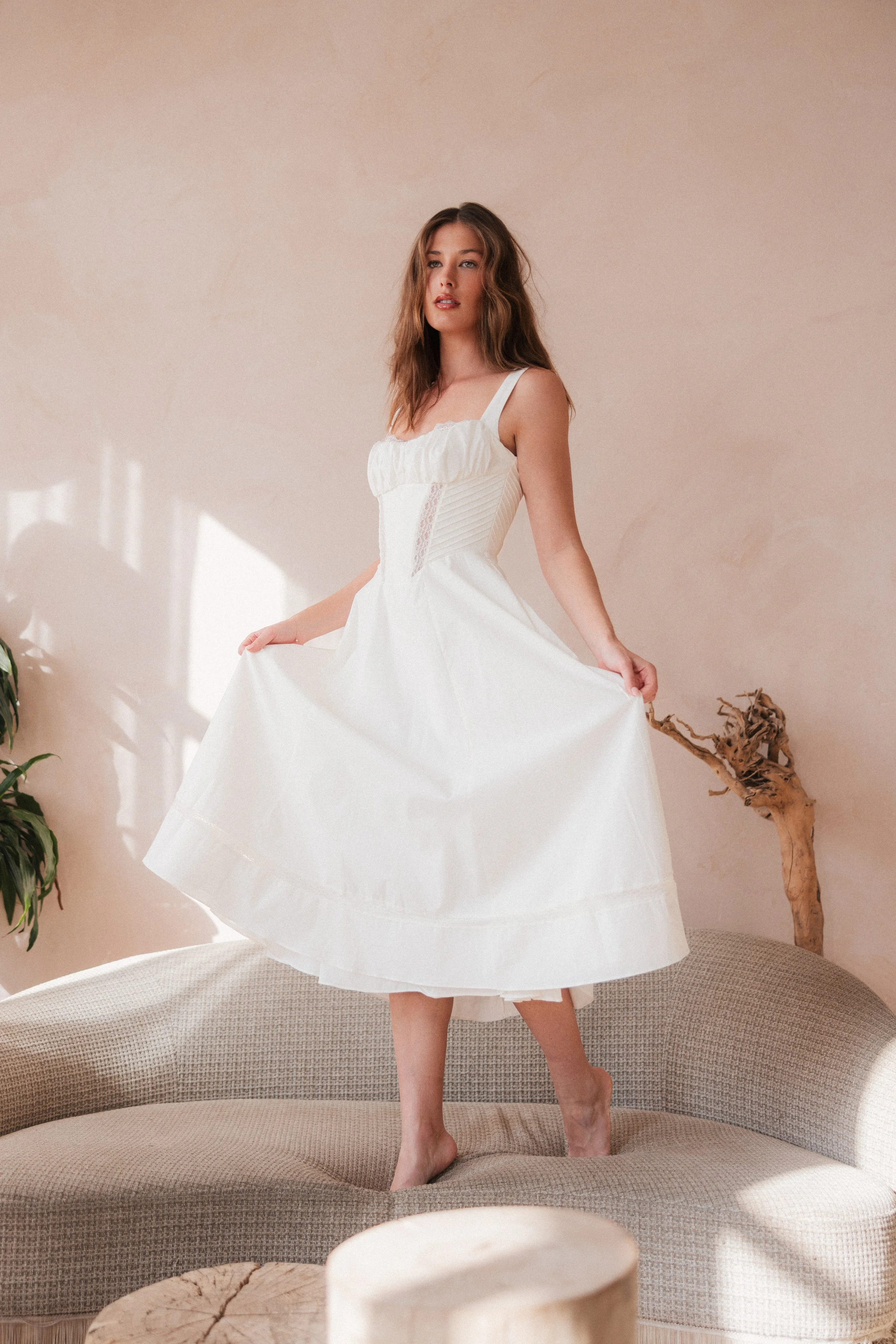 A woman in a white dress standing on a beige sofa, holding the hem of her dress, with minimalist decor including a twisted wood sculpture and potted plant in a neutral room.