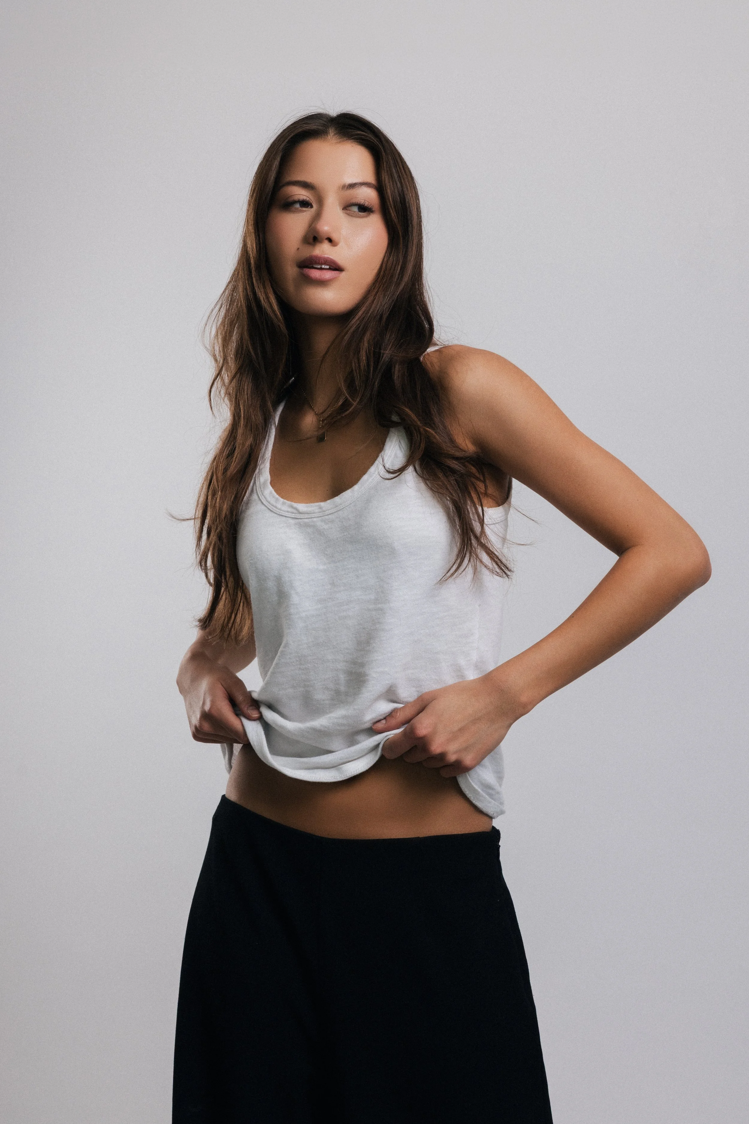 A young woman with long brown hair wearing a white tank top and black pants, standing against a plain light gray background.