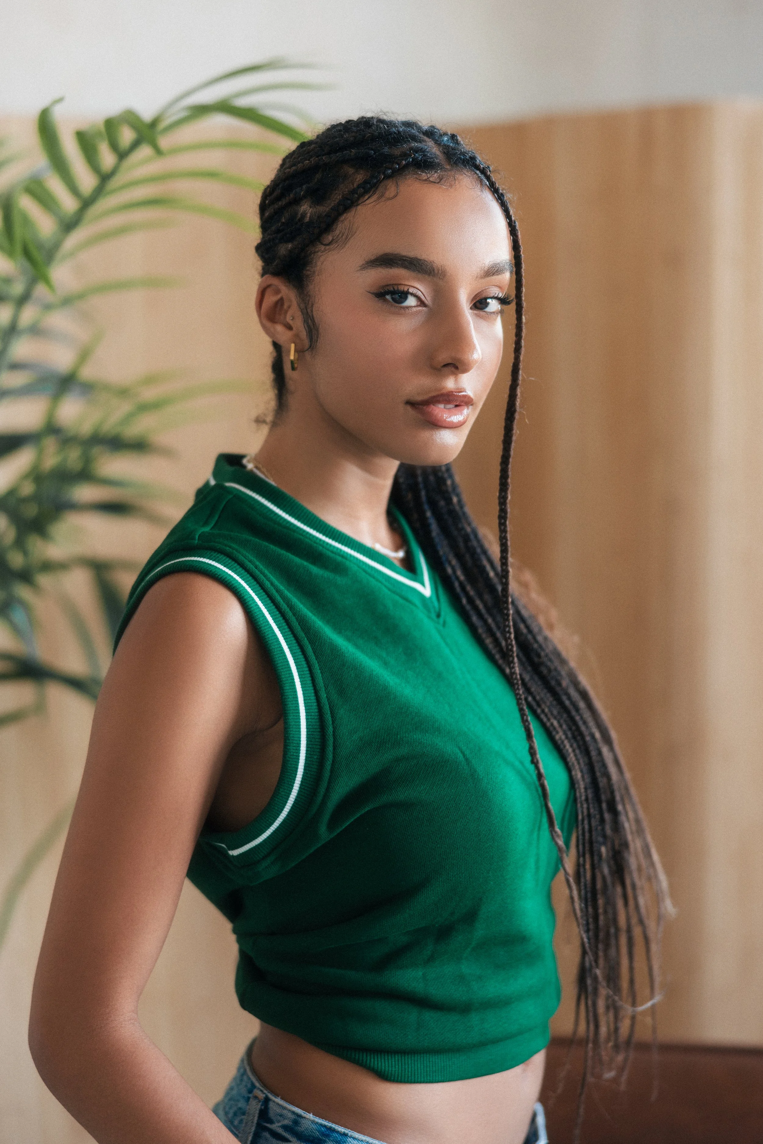 A young woman with medium brown skin and dark braided hair wearing a green sleeveless athletic top and denim shorts, posing indoors near a plant with large green leaves.