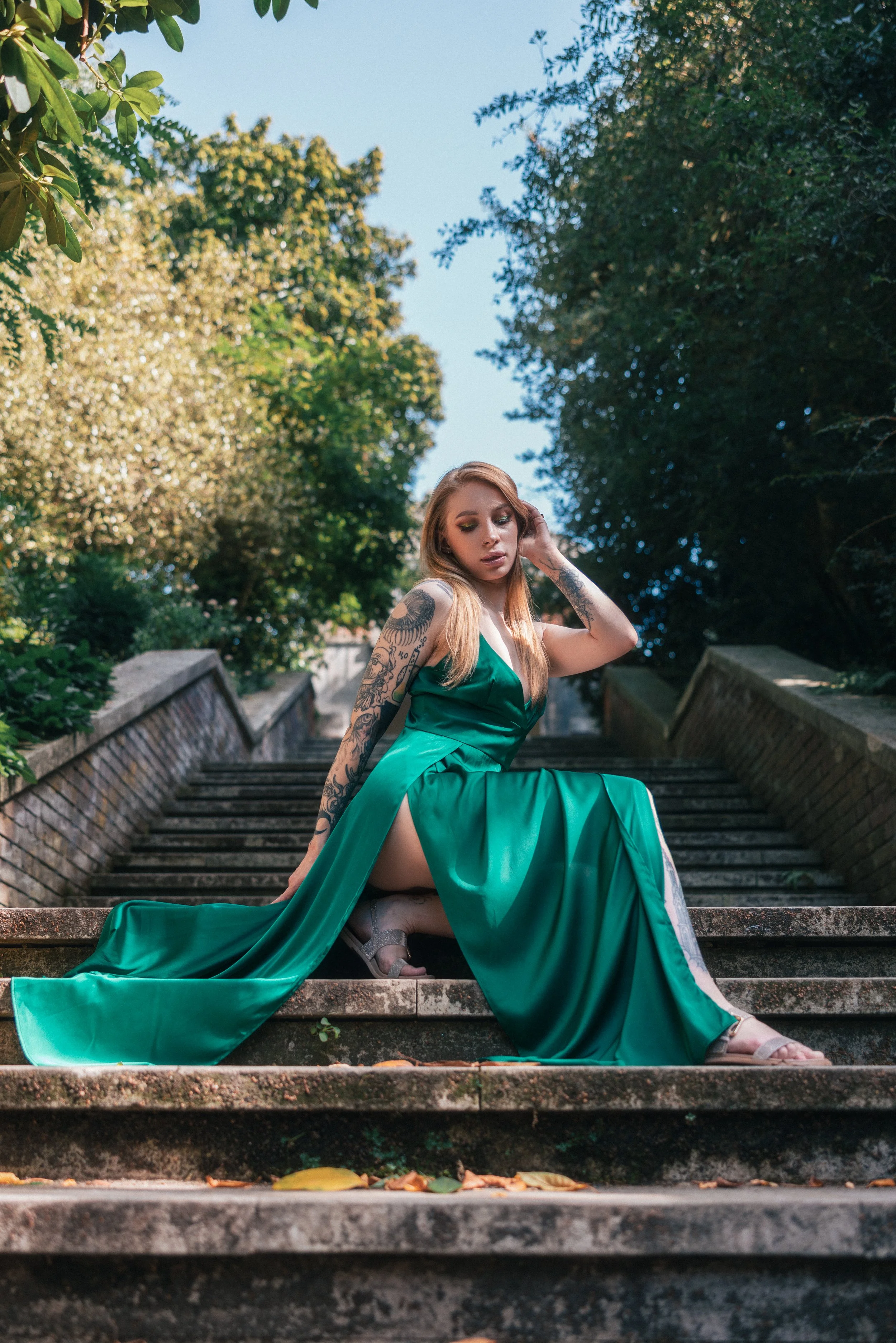 A woman with tattoos wearing a green dress sitting on outdoor steps surrounded by trees.