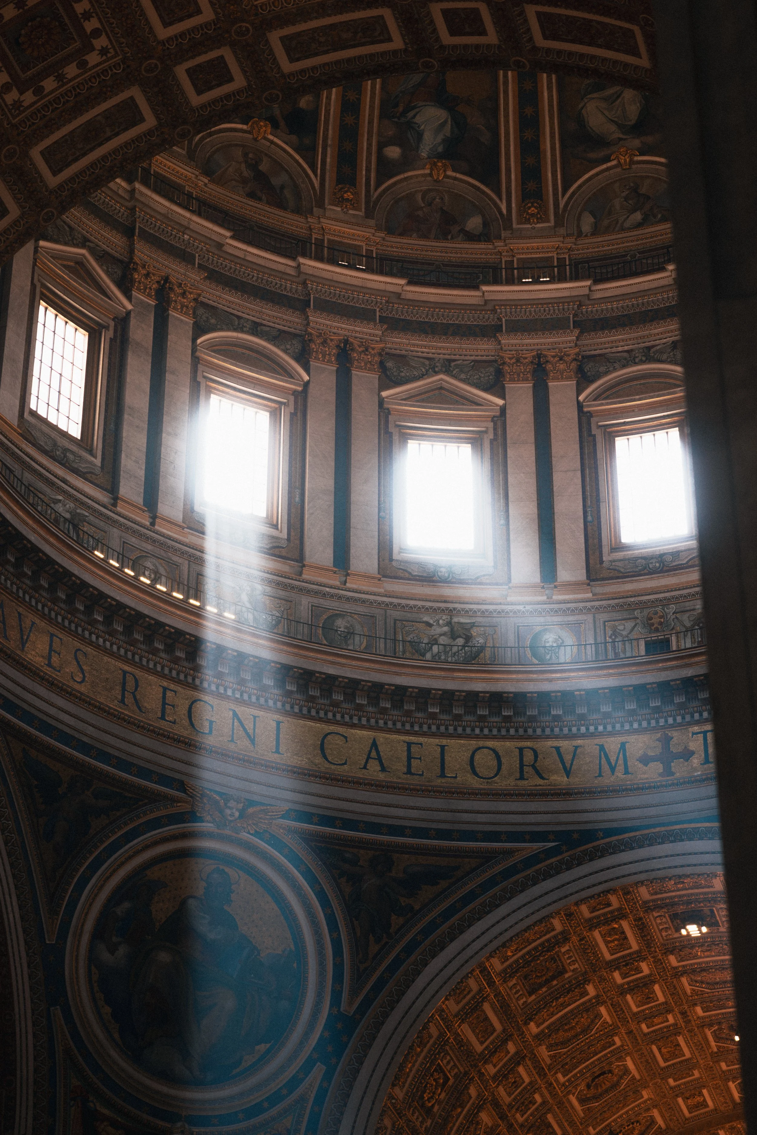 Interior view of a grand domed building with ornate decorations, large windows letting in sunlight, and Latin inscriptions on the walls.