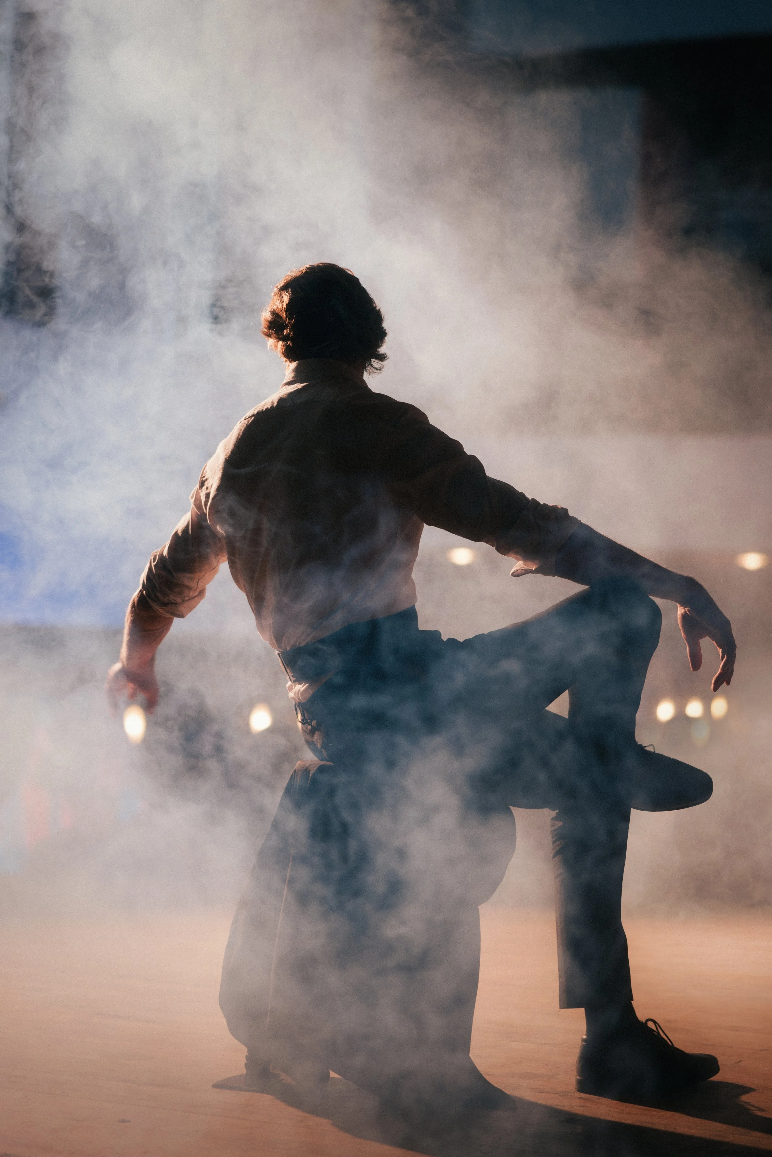 A person with medium-length hair, kneeling with one knee on the ground and arms spread wide, surrounded by fog or smoke, with a dramatic lighting creating a silhouette effect.