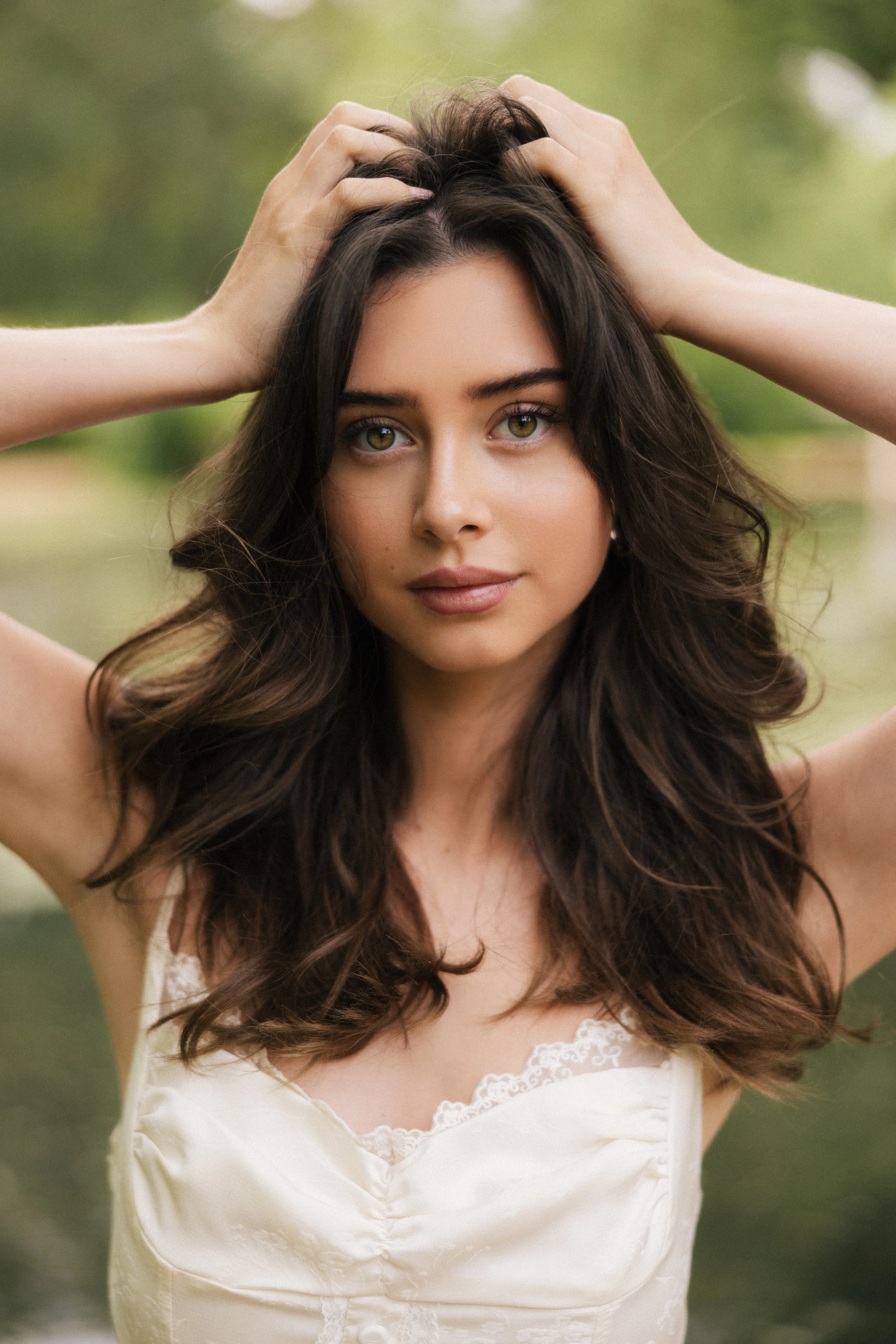A young woman with long wavy brown hair and light green eyes, wearing a white dress with lace details on the neckline, standing outdoors in a natural setting with blurred green background, holding her hair back with both hands.