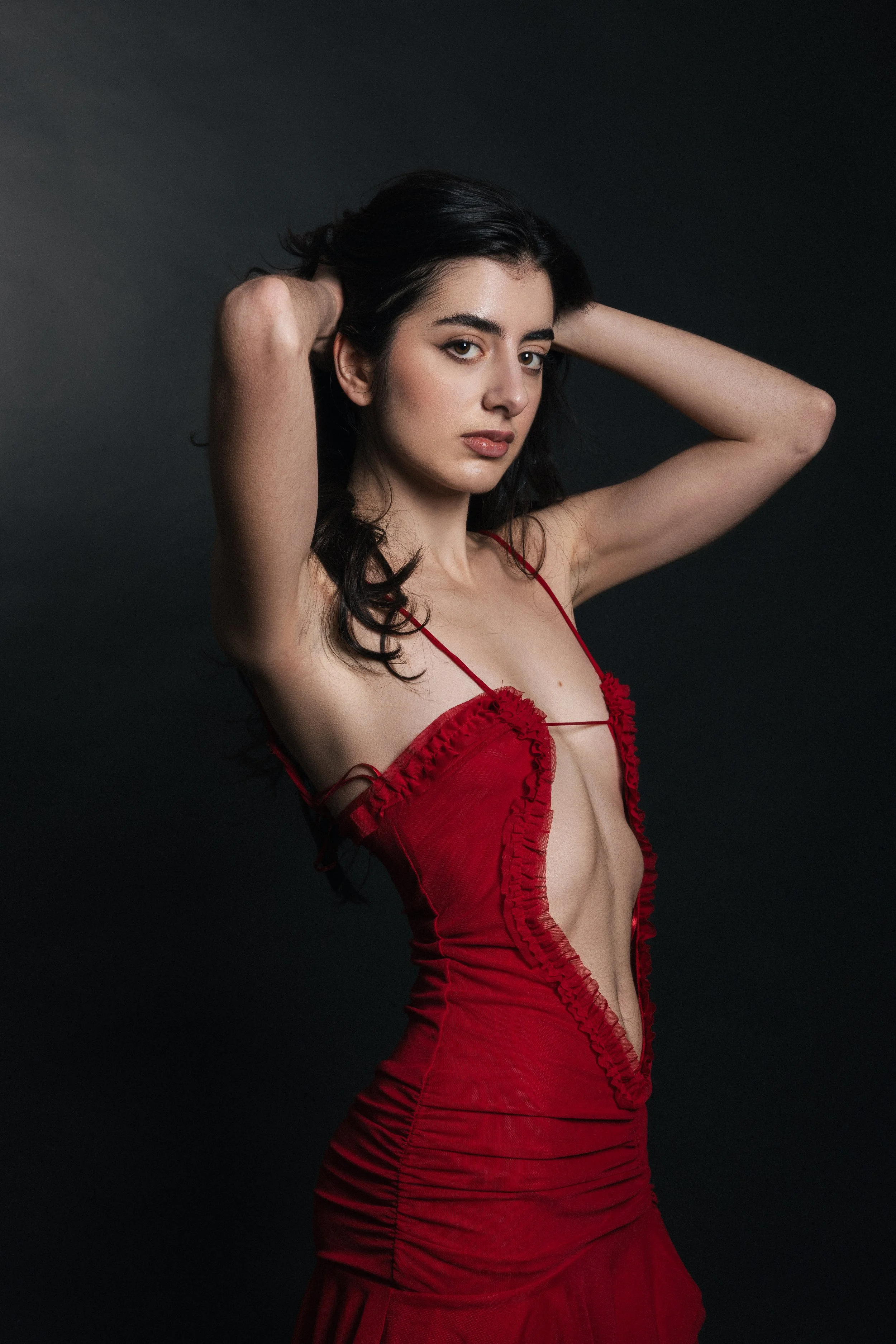 A woman in a red dress with ruffled details poses with her arms raised, dark hair, and a neutral expression against a black background.