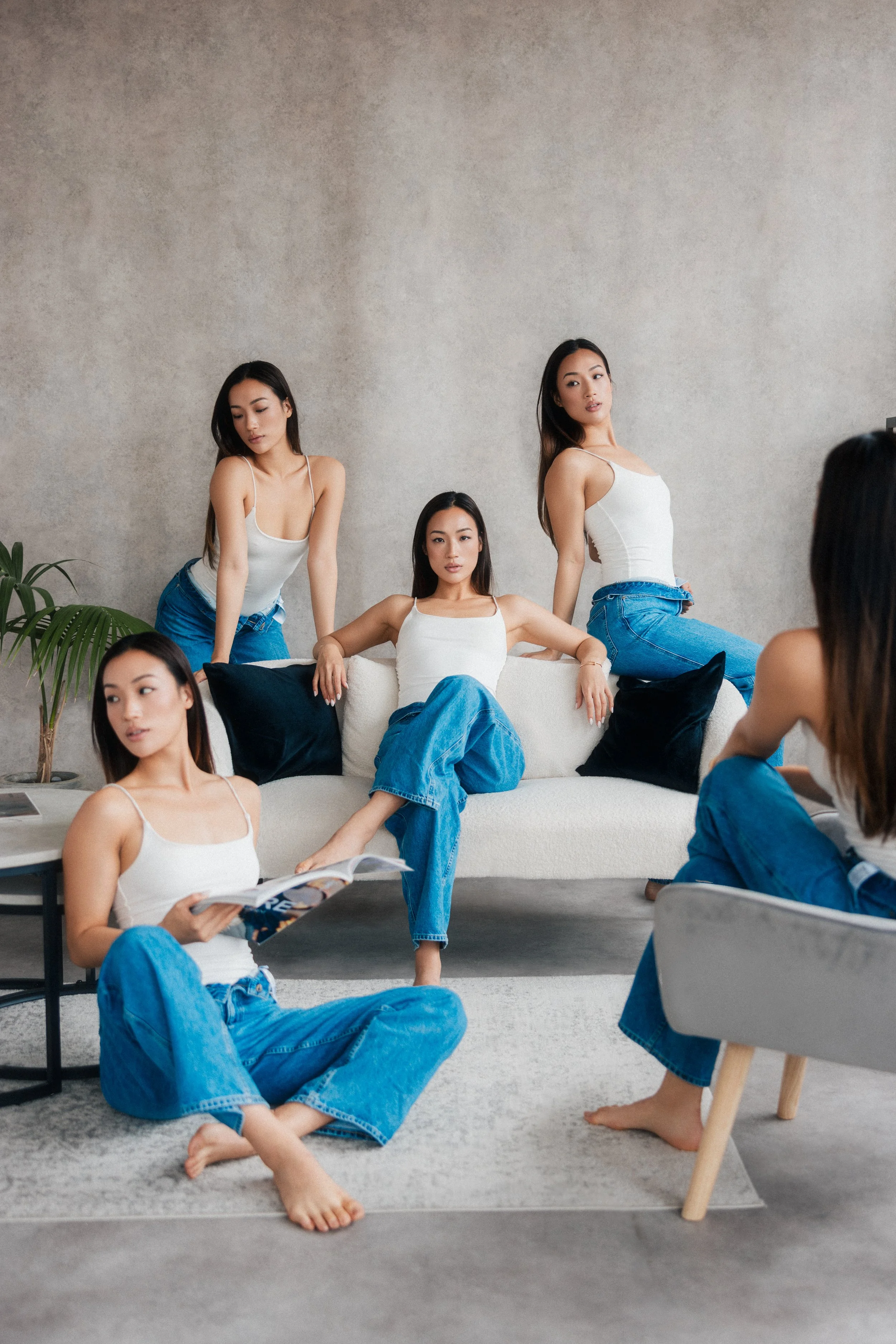 Six young women with light to medium skin tones and long dark hair, wearing white tank tops and blue jeans, are positioned around a modern living room. Two women are sitting on a gray sofa with black pillows, one is seated on the floor reading a maga