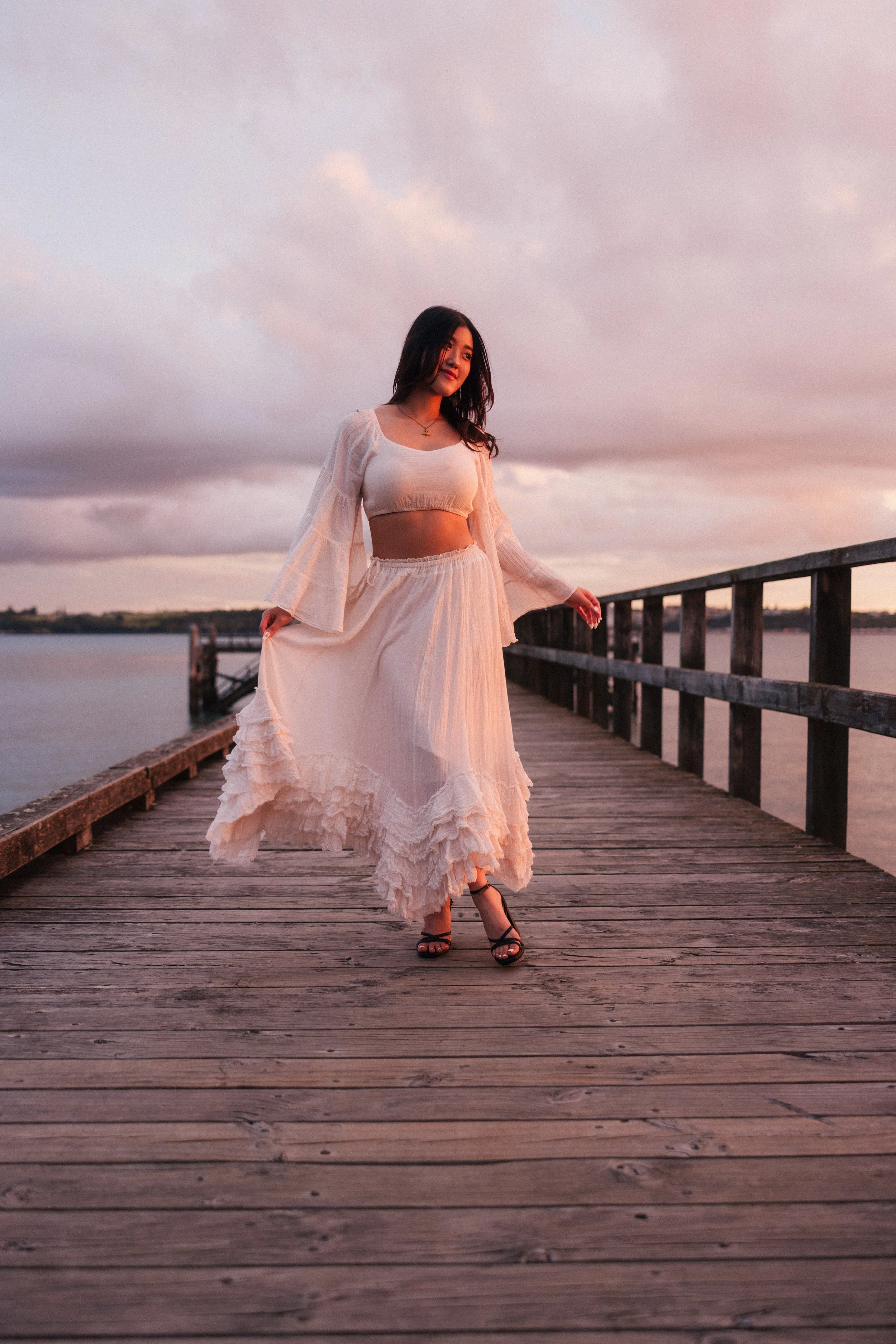 A woman standing on a wooden dock at sunset, wearing a white crop top and flowing skirt, with a cloudy sky in the background.