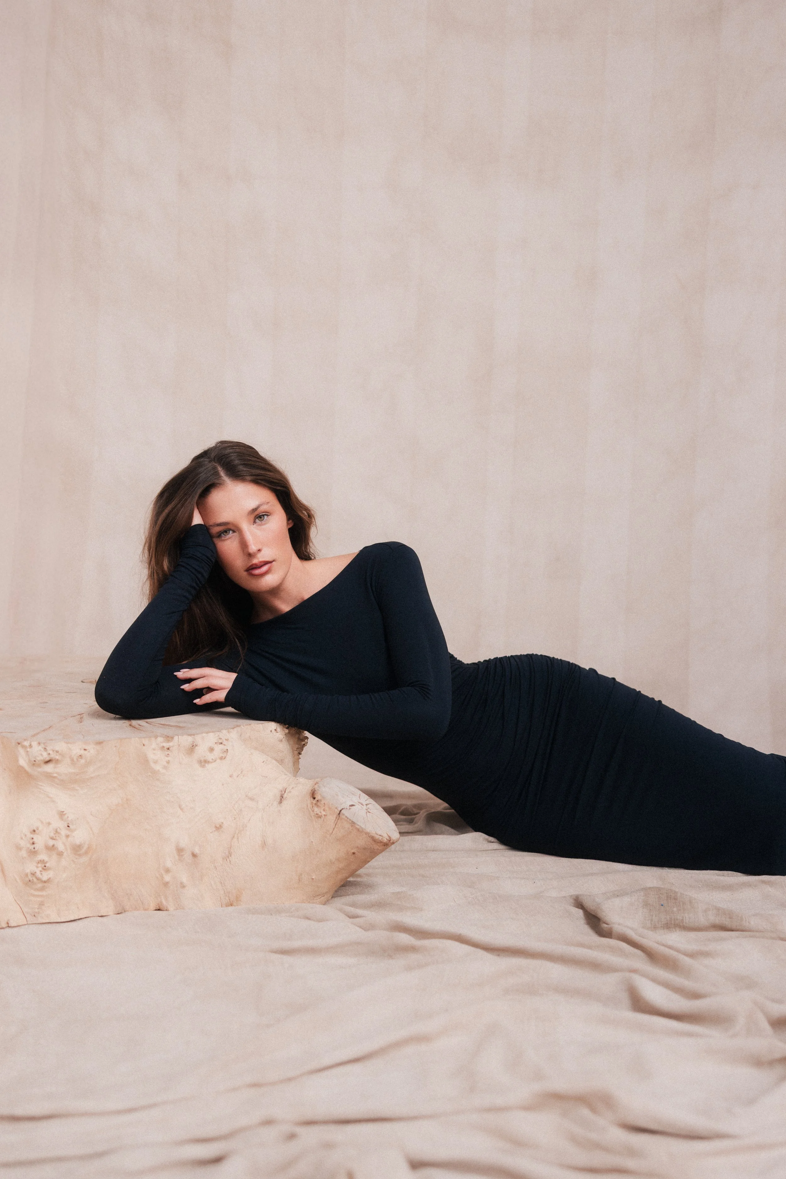 A woman lying on her side on a beige fabric surface, resting her head on her hand, in a black long-sleeve dress against a plain beige wall background.