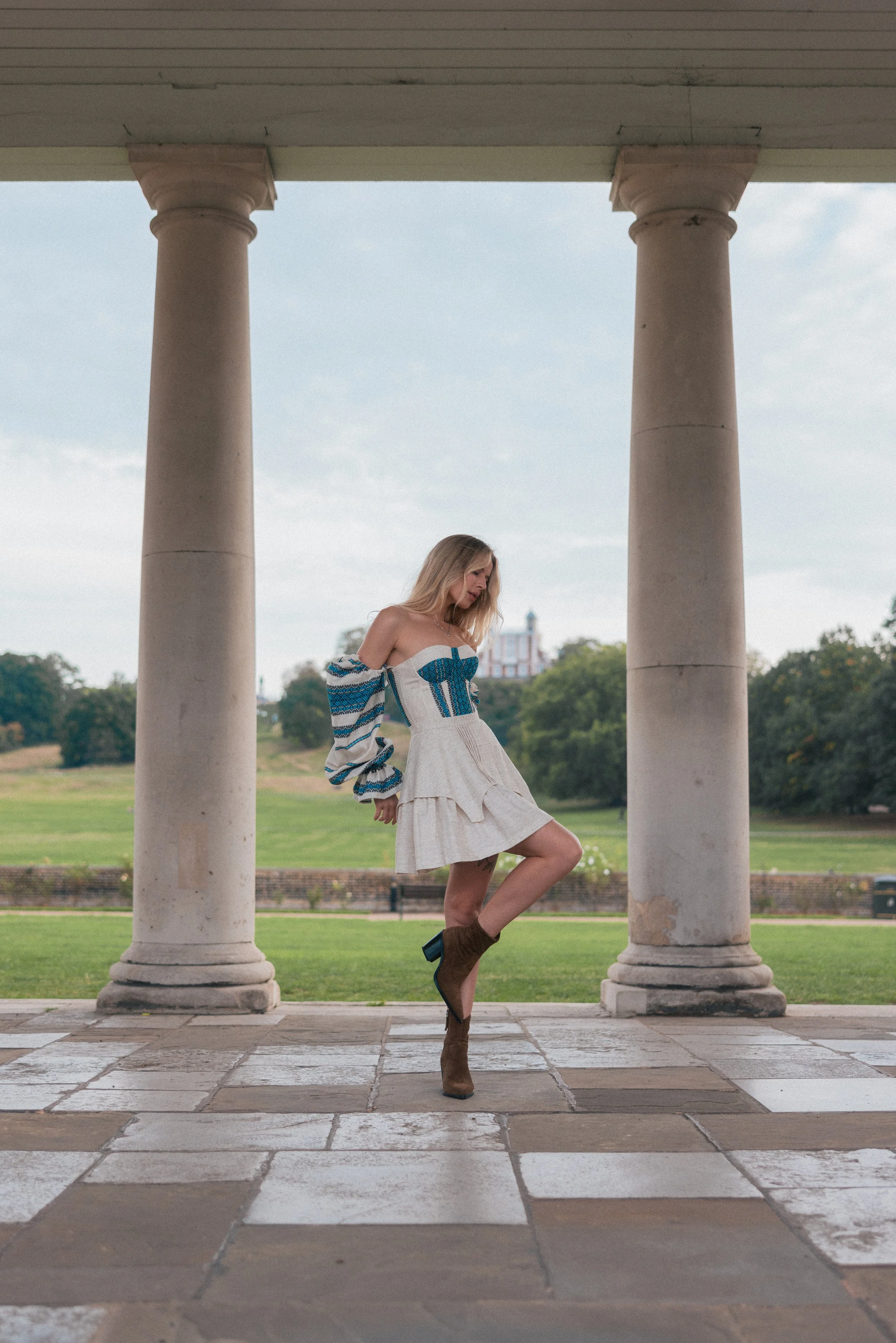 A young woman with long blonde hair poses outdoors under white columns, wearing a white skirt, a blue and white patterned off-the-shoulder top, and brown high-heeled boots, with a landscape of trees and a building in the background.