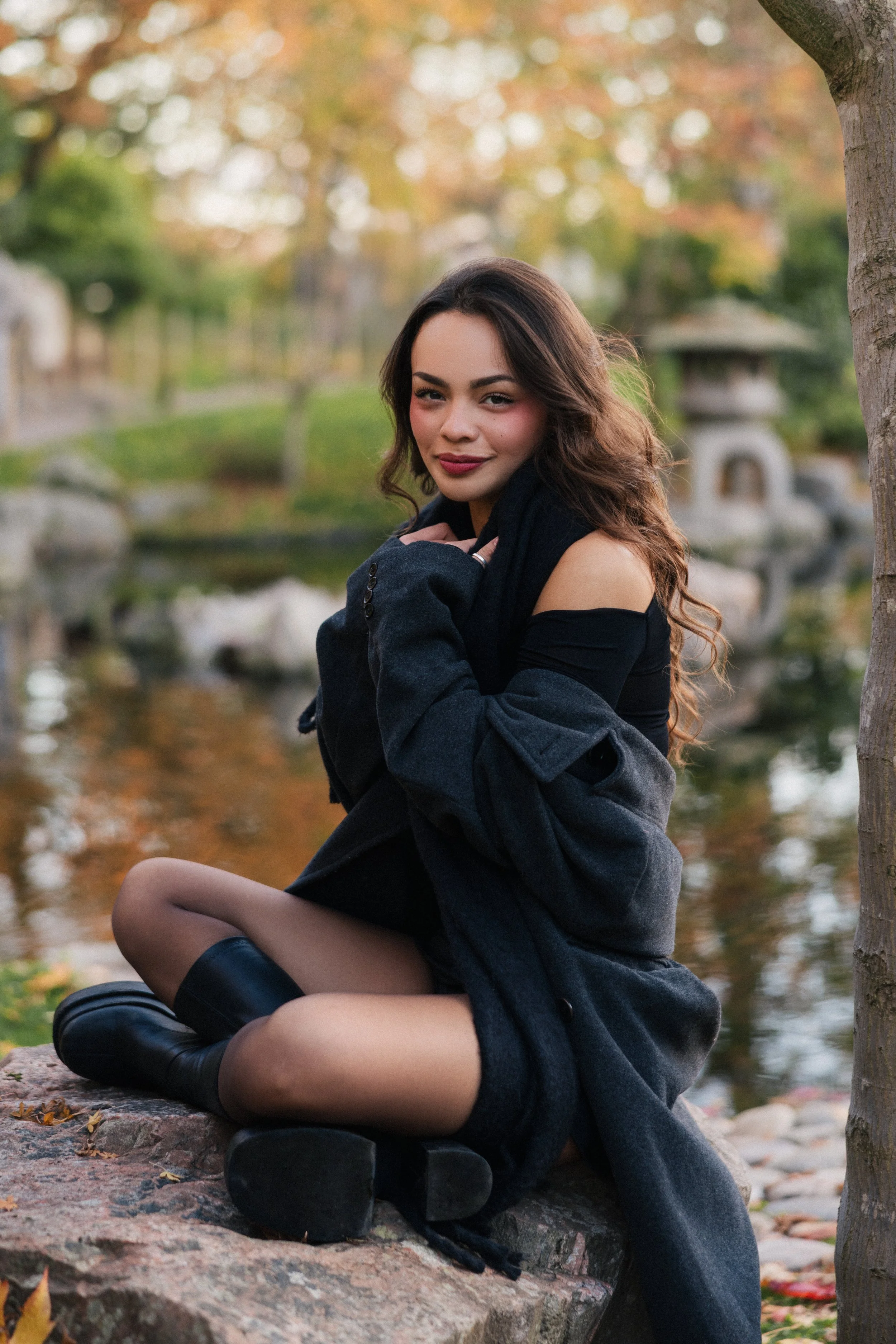 A young woman with wavy brown hair sitting on a rock by a pond during autumn, wearing a black off-shoulder top, black boots, and a dark coat, with trees and fall foliage in the background.