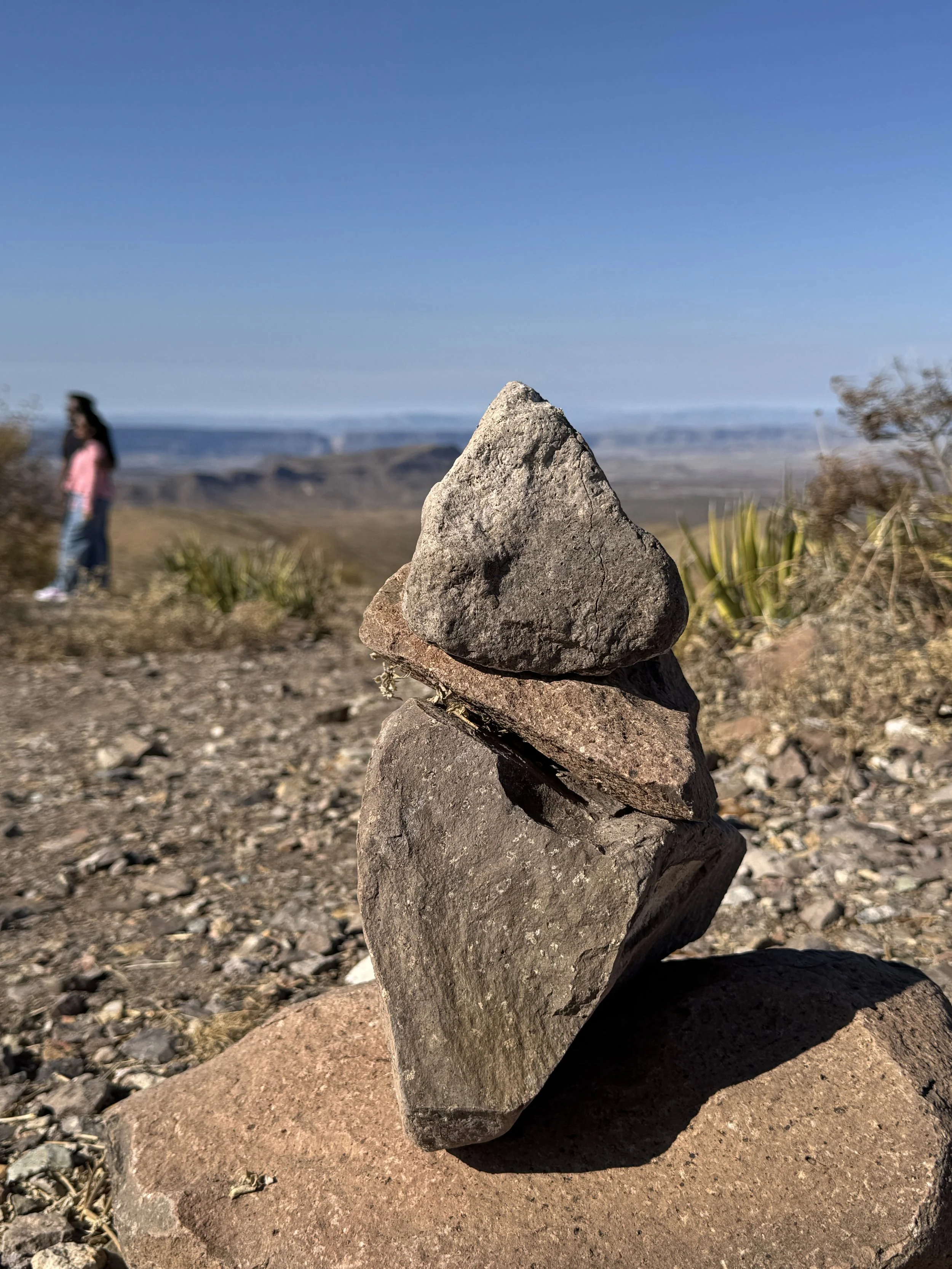 A stack of rocks balanced one on top of the other in an outdoor desert landscape with a blue sky and distant canyon in the background. There is a blurred person in a pink jacket and blue jeans standing to the left.