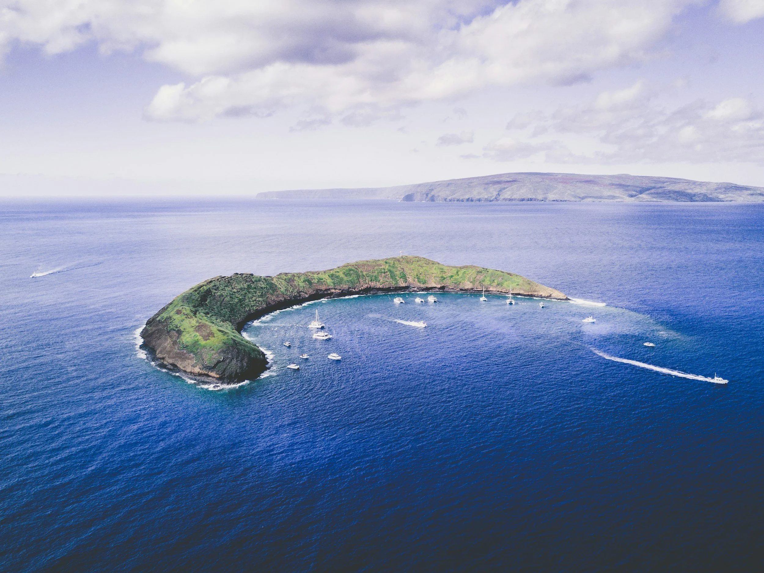A large, circular, green island with a harbor and boats, surrounded by deep blue ocean and distant landforms under a partly cloudy sky.