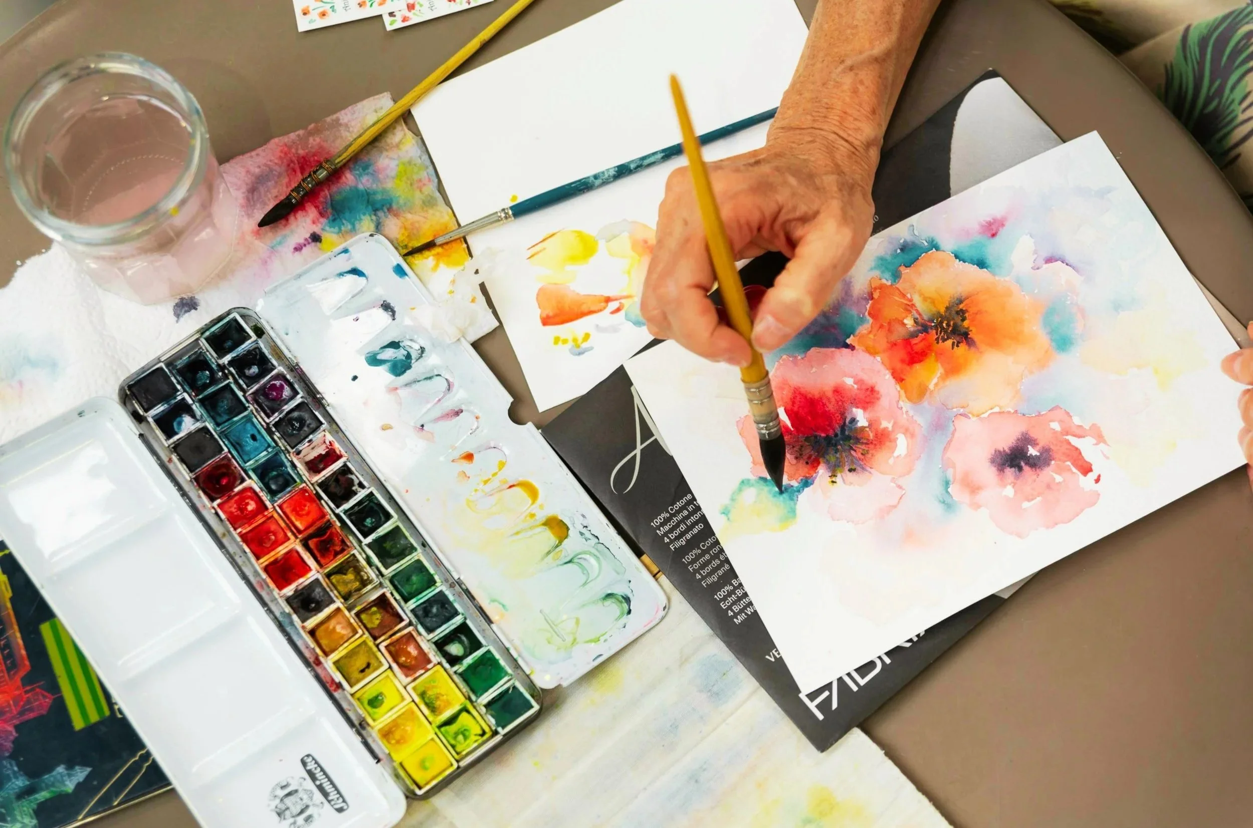Close-up of an artist's workspace showing watercolor paints, paintbrushes, a glass of water, and a watercolor painting of colorful flowers on paper.