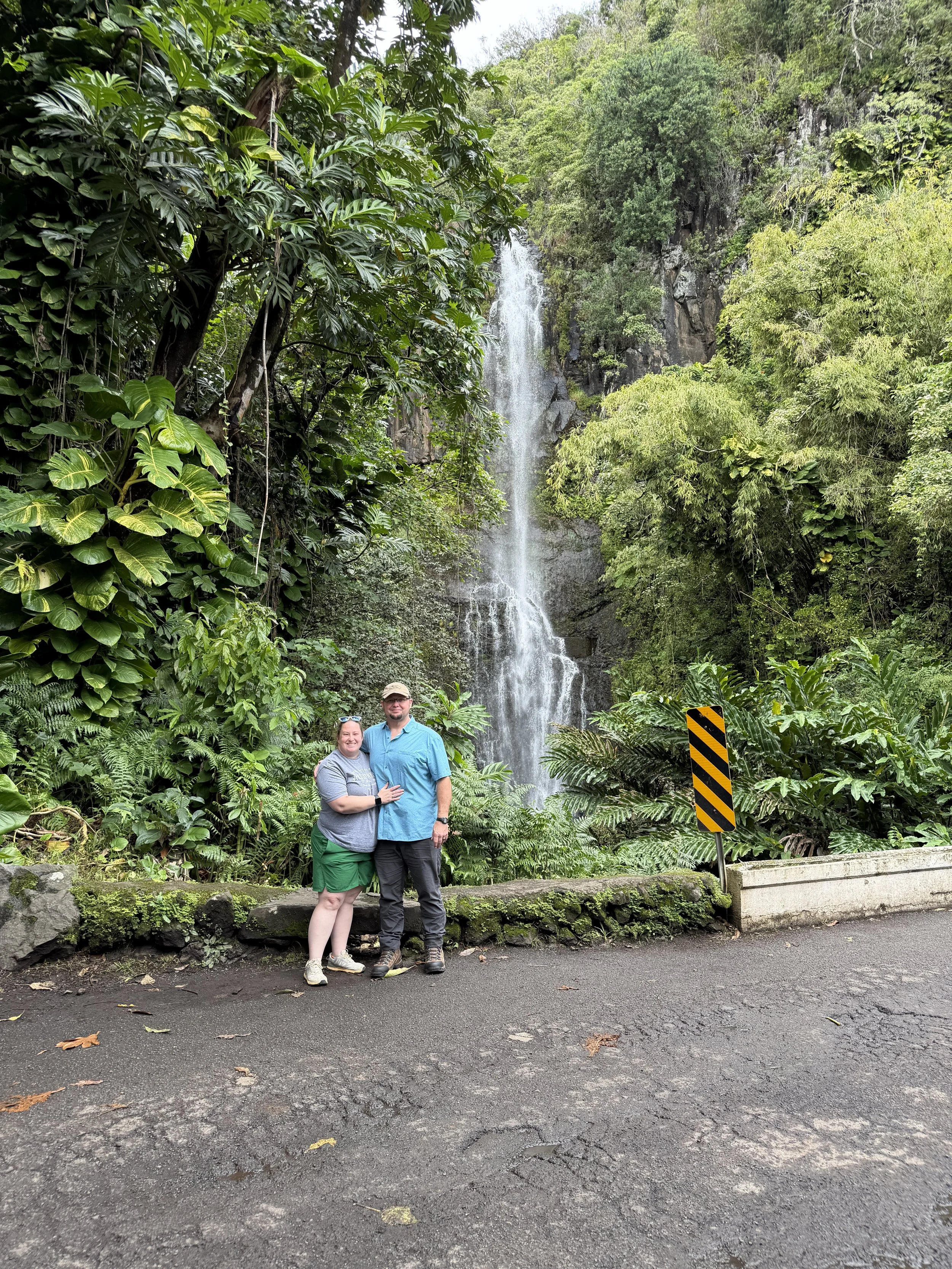 A couple stands in front of a waterfall surrounded by lush greenery in a forest. The woman is wearing a gray shirt and green shorts, and the man is wearing a blue shirt and black pants. They are smiling and standing close together.