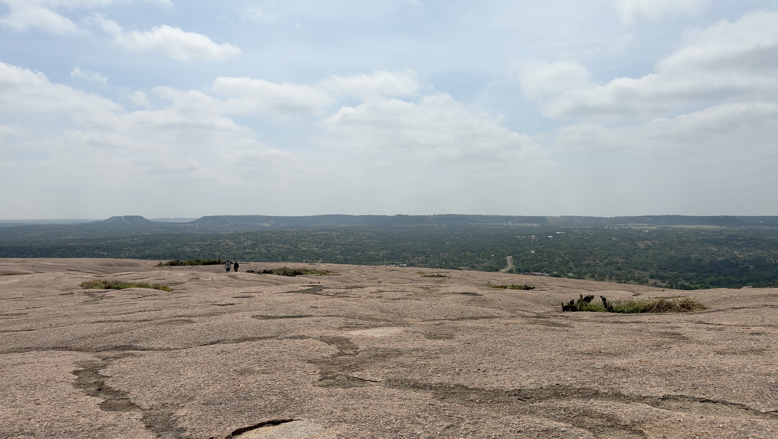 A vast rocky landscape with two people walking in the distance, overlooking a green valley with hills and a cloudy sky.
