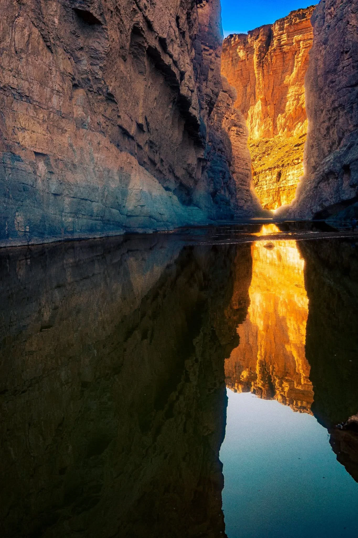 A canyon with steep rocky walls illuminated by sunlight, and a calm river flowing at the bottom reflecting the canyon walls and the blue sky.