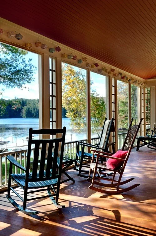 Sunlit porch with wooden rocking chairs overlooking a lake and trees.
