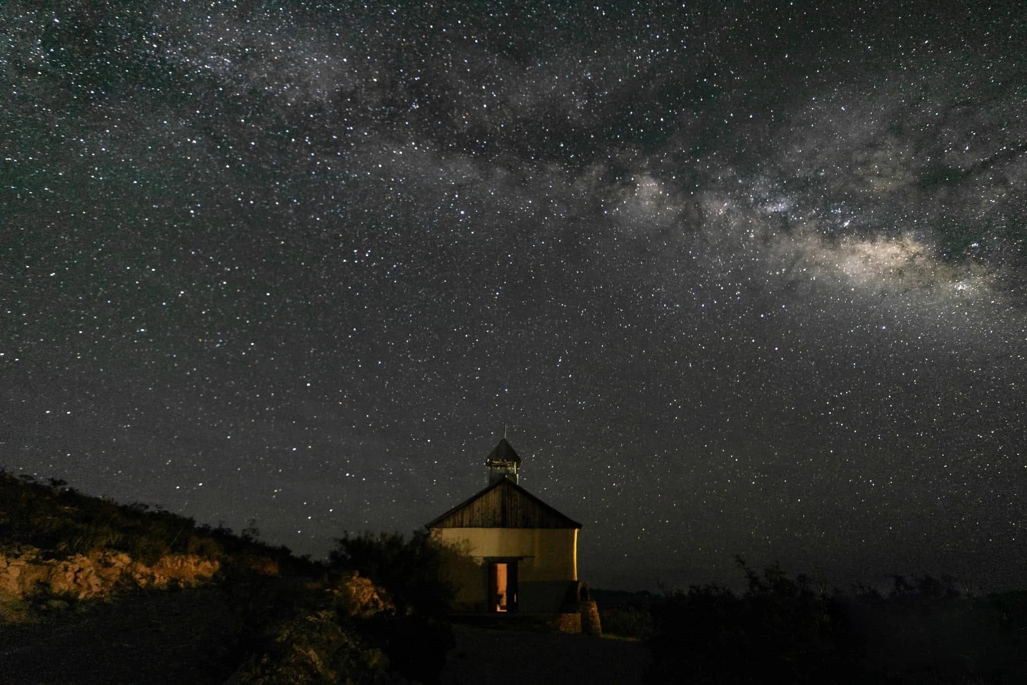 Night sky filled with stars and the Milky Way galaxy, with a small house or building with a pointed roof in the foreground.